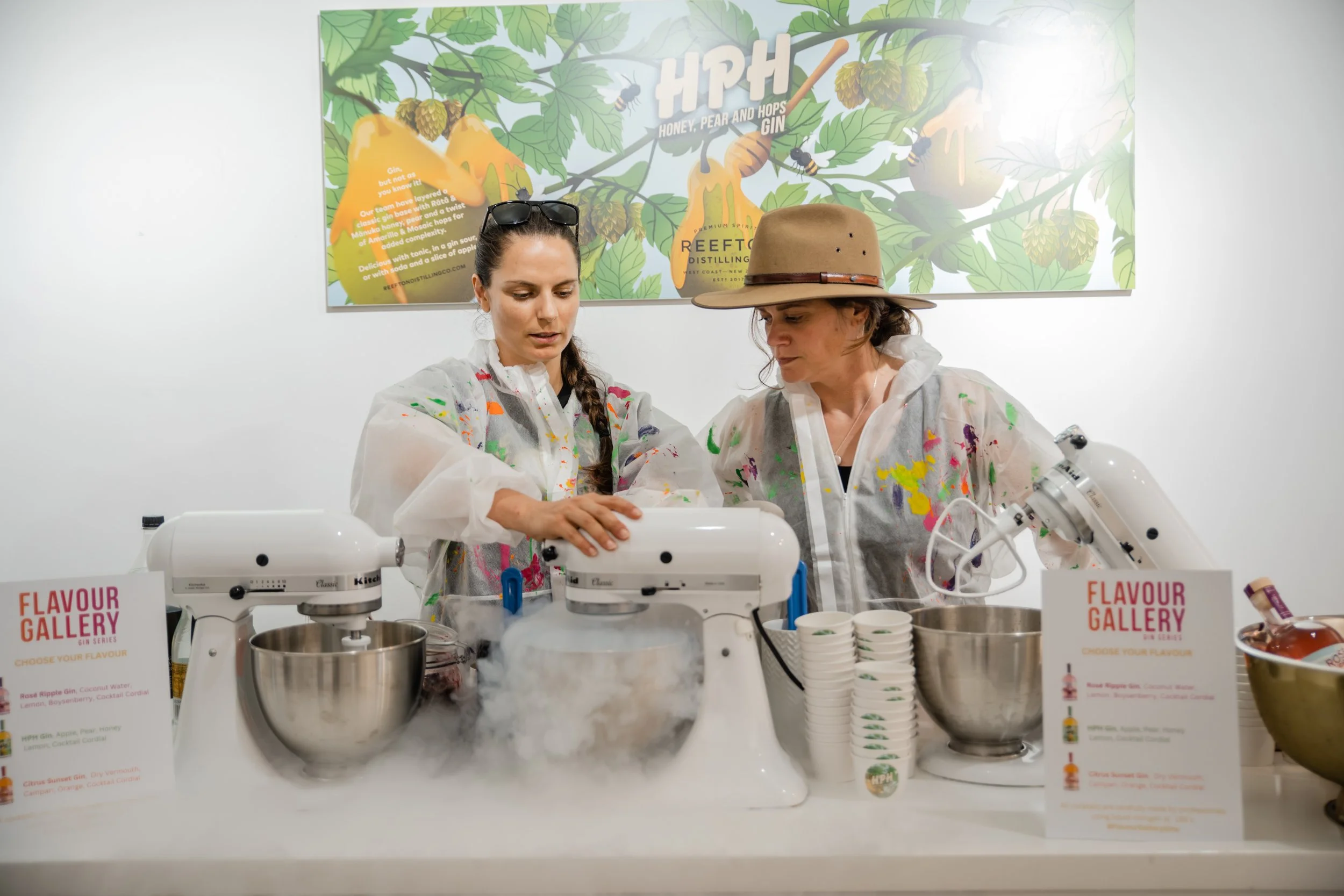 Two women in protective jackets working with mixing machines surrounded by cups and flavor signs at a gin tasting event, with a colorful botanical-themed poster in the background.