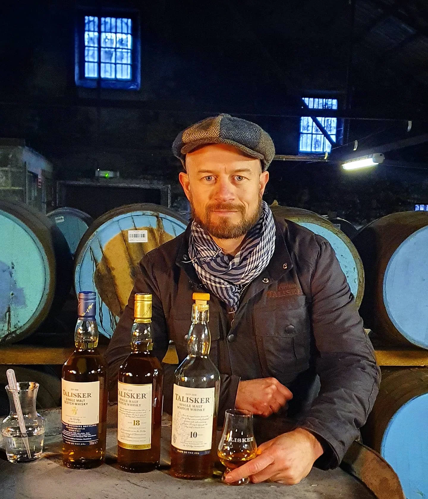 A man sitting at a table in a distillery, surrounded by barrels, holding a glass of whisky, with three bottles of Talisker single malt whisky in front of him.
