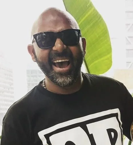 A man with a shaved head, beard, and sunglasses, smiling widely, wearing a black T-shirt with a graphic design, standing outdoors with a large green leaf and city buildings in the background.