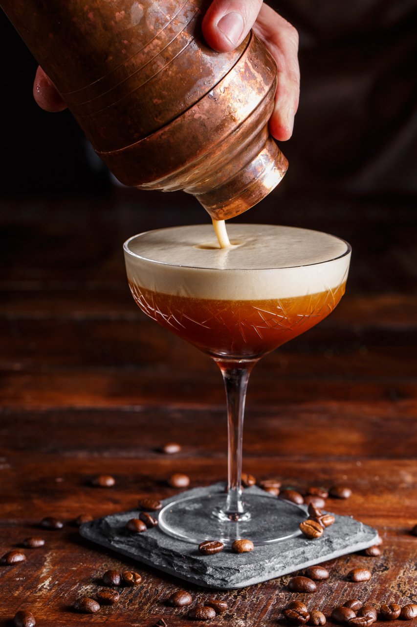 A bartender pours coffee liqueur into a cocktail glass filled with a dark, layered coffee-based cocktail, with coffee beans scattered on a wooden surface.