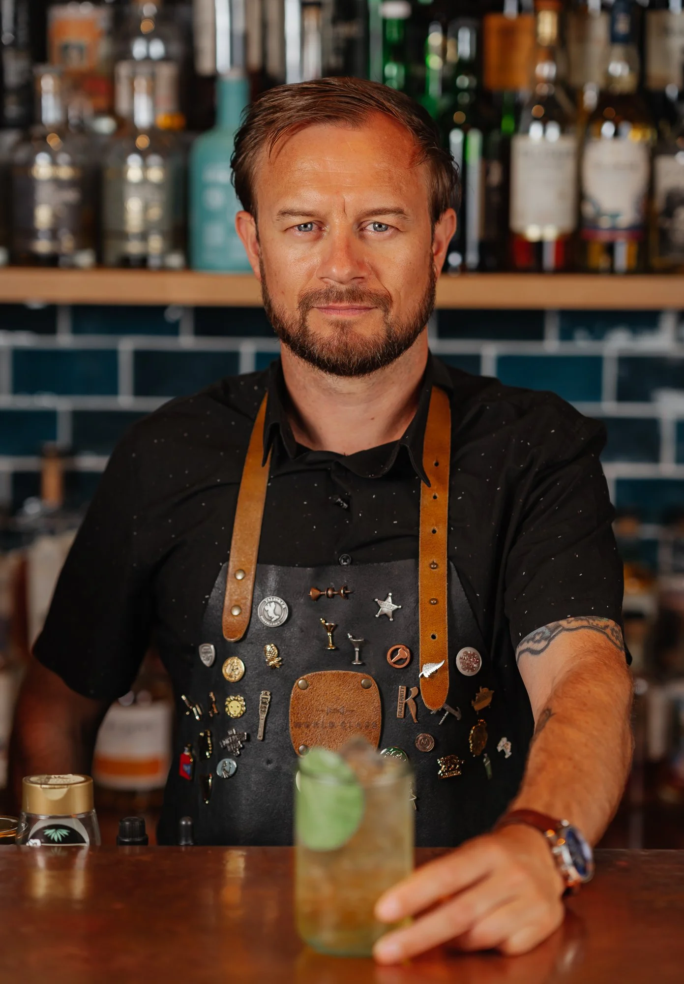 A bartender with a beard and tattoos, wearing a black shirt and pin-covered apron, prepares a cocktail behind a bar.