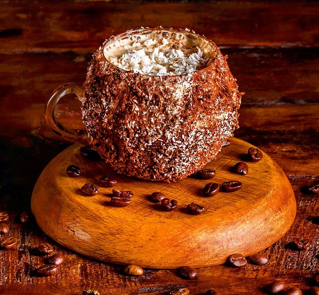A coffee mug wrapped in chocolate with shredded coconut on top, placed on a wooden round board with scattered coffee beans around it.