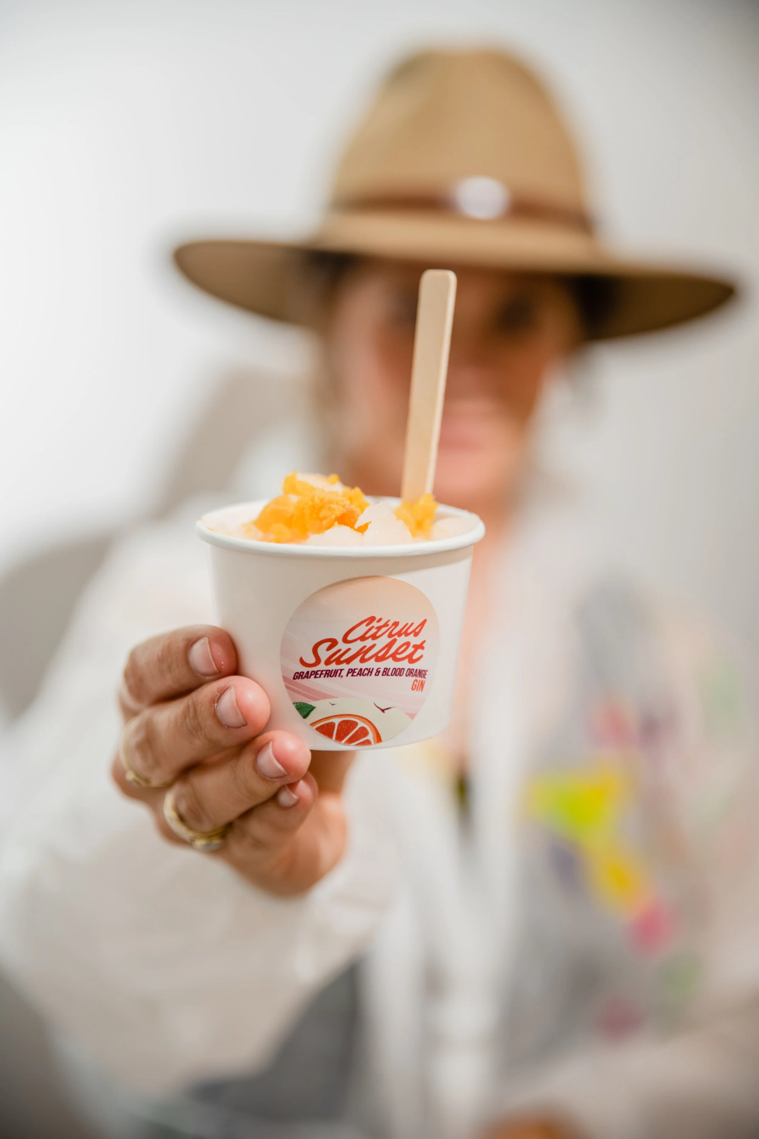 Person holding a cup of citrus sunset ice cream with a blurred background that includes a woman wearing a wide-brimmed hat.
