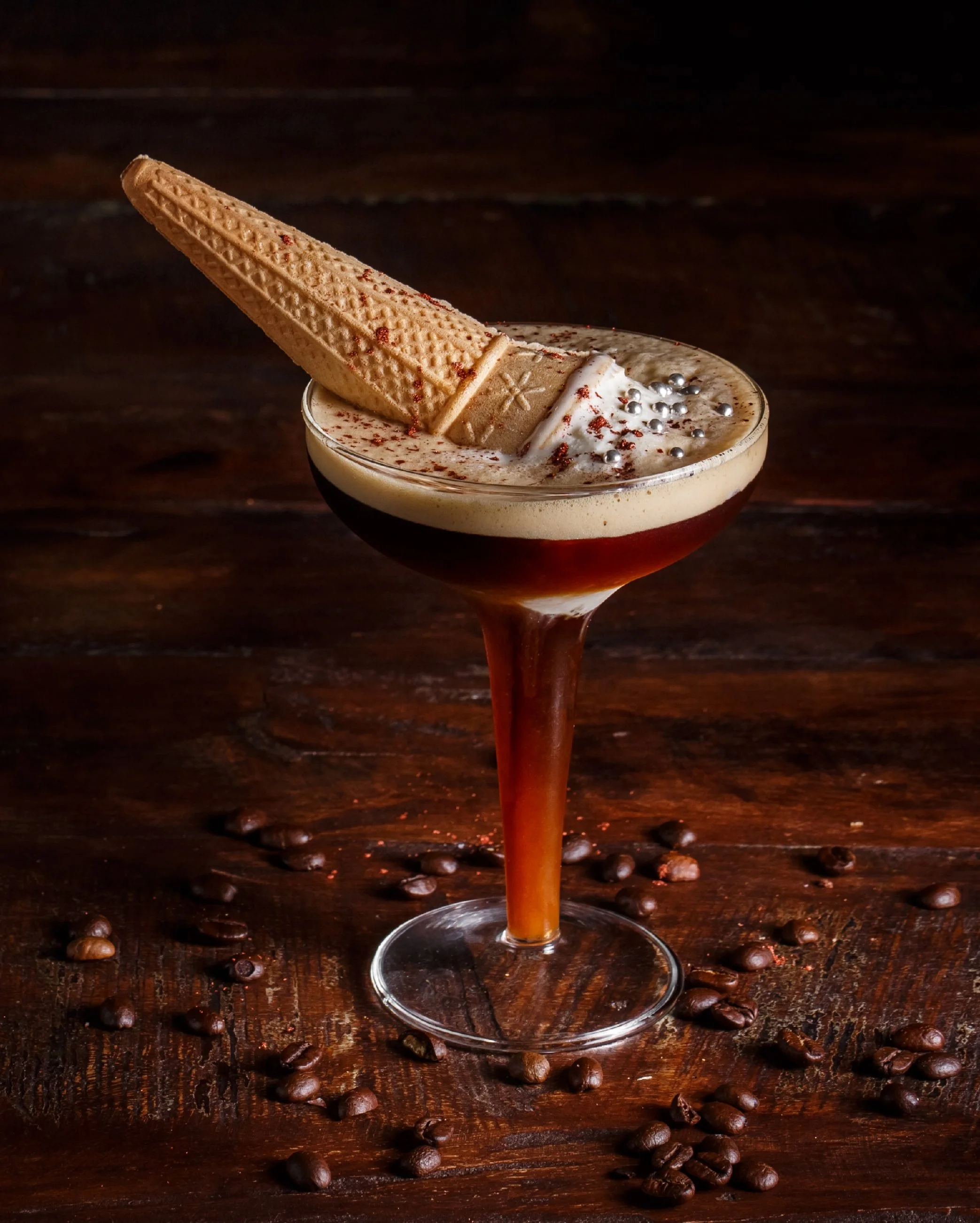 A coffee cocktail in a glass with a wafer and silver sprinkles on top, surrounded by scattered coffee beans on a dark wooden surface.