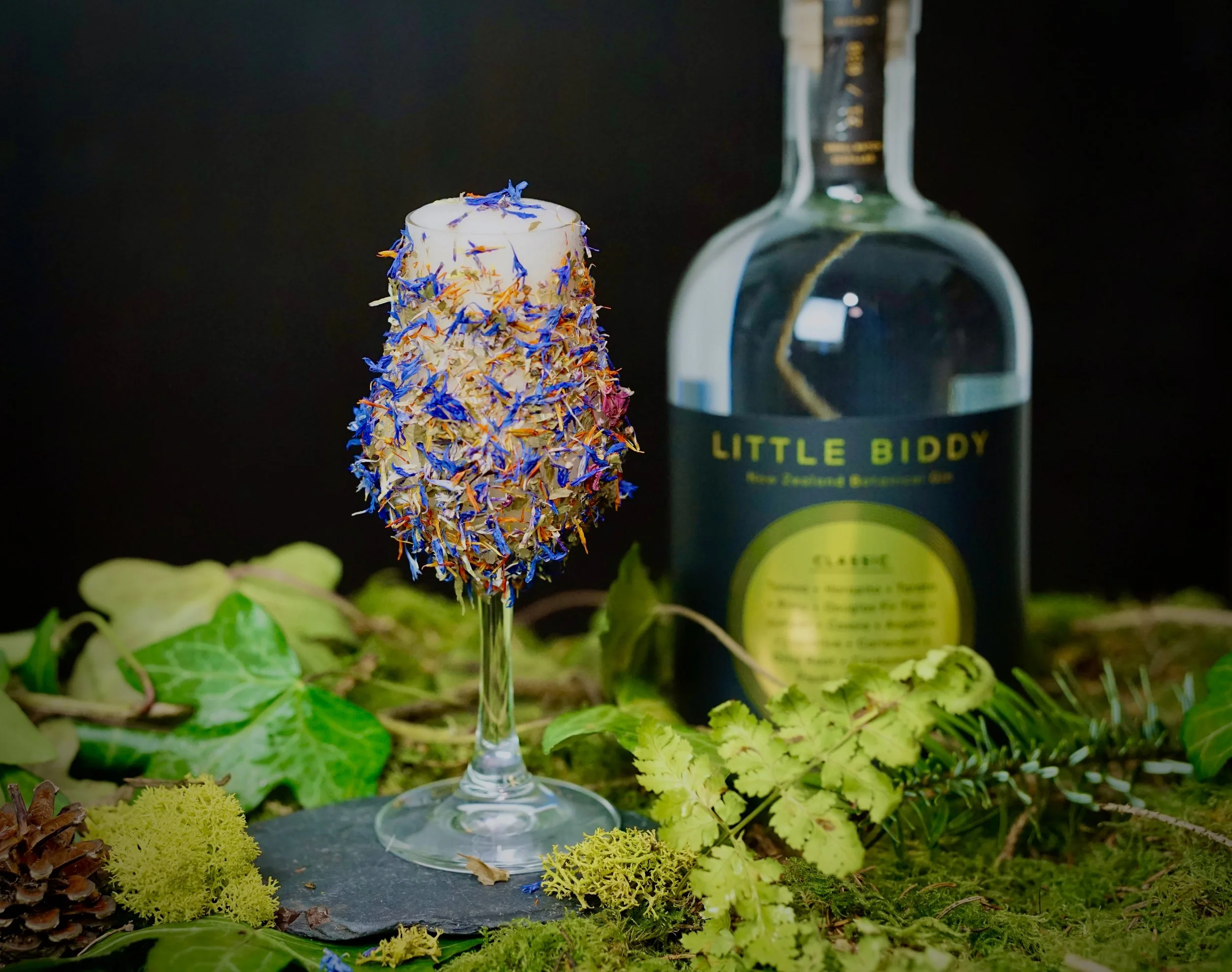 A glass filled with dried blueberry and flower petals, with a bottle labeled 'Little Biddy' behind it, set on a bed of moss and green leaves.