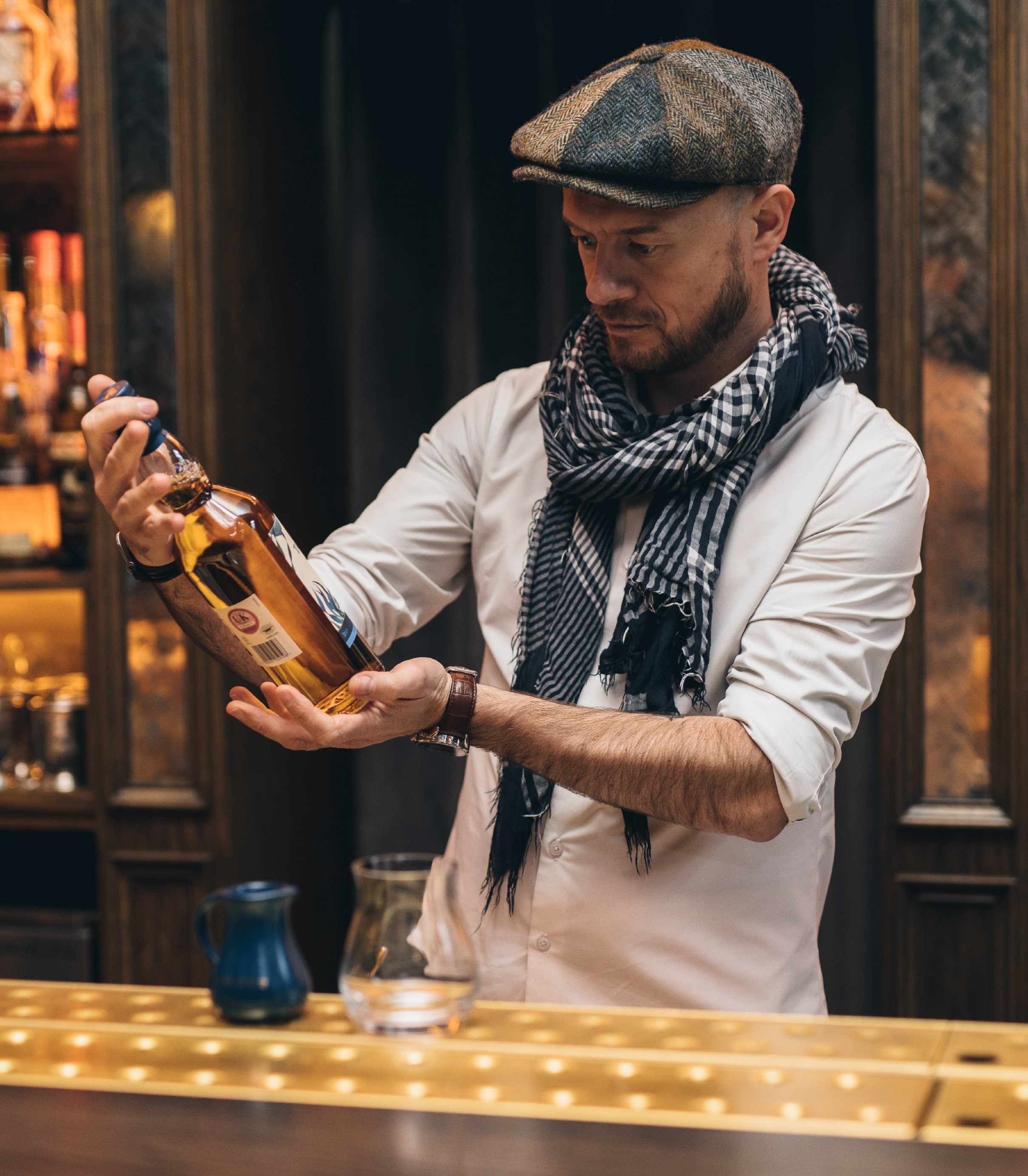 A man in a flat cap, white shirt with rolled-up sleeves, and a checkered scarf pours a drink from a bottle into a glass behind a bar.