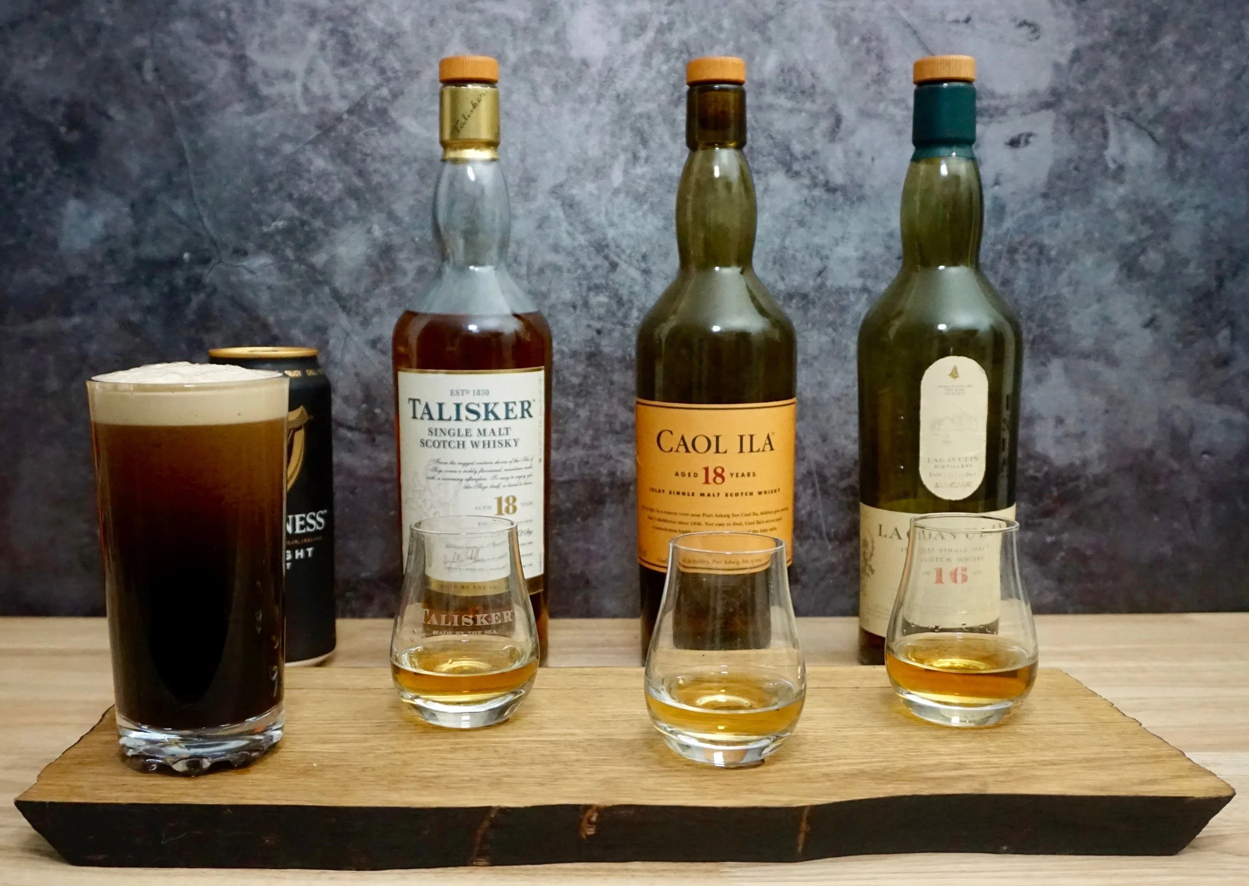Three bottles of whisky, one dark stout beer, three whisky glasses with whisky, on a wooden tray against a dark gray background.