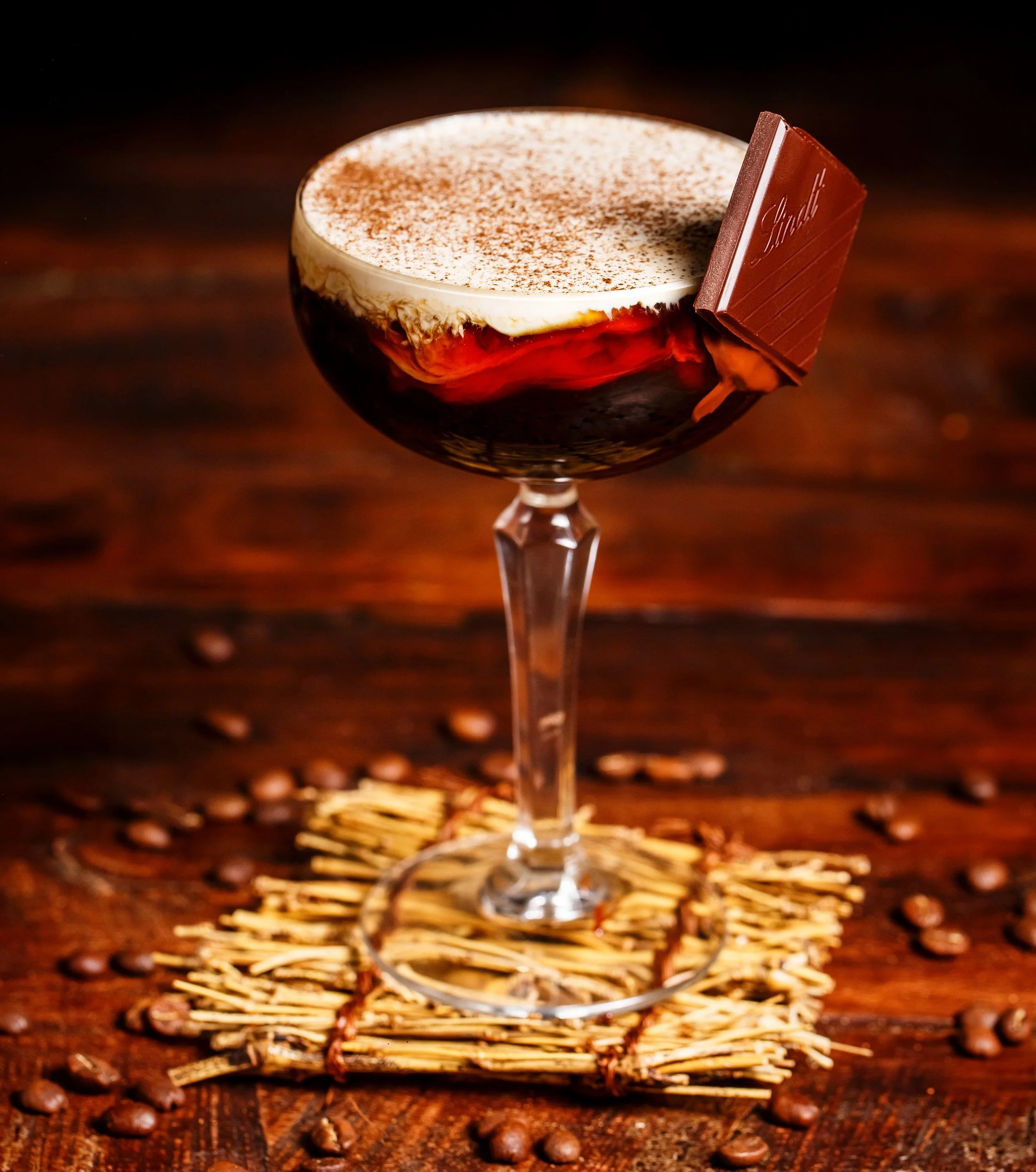 A cocktail in a stemmed glass with whipped cream, cocoa powder, and a piece of chocolate on the rim, set on a wooden surface with coffee beans around it.