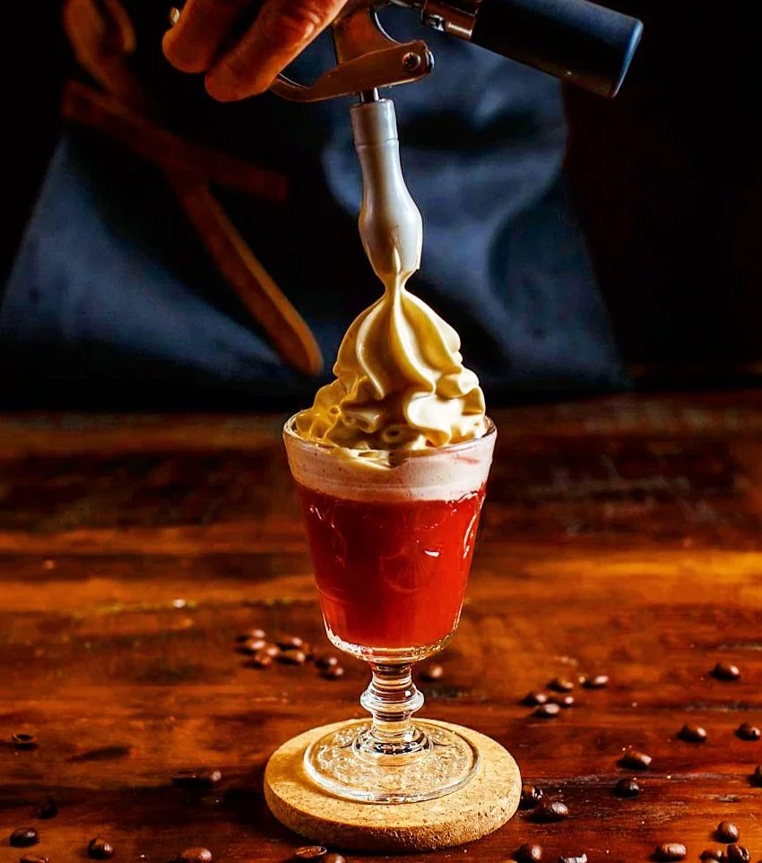 A coffee beverage in a glass with a dark red liquid at the bottom, topped with a swirl of vanilla soft serve ice cream, being decorated with a siphon dispenser.