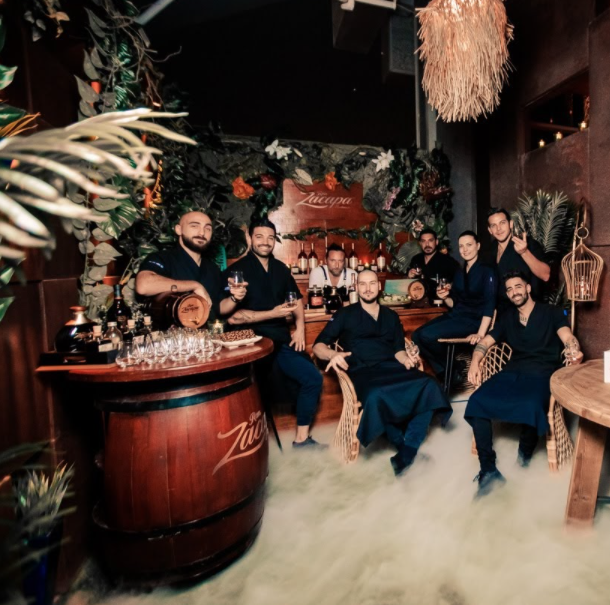 Group of people in a bar or restaurant setting with tropical decorations, holding drinks, and smiling at the camera.