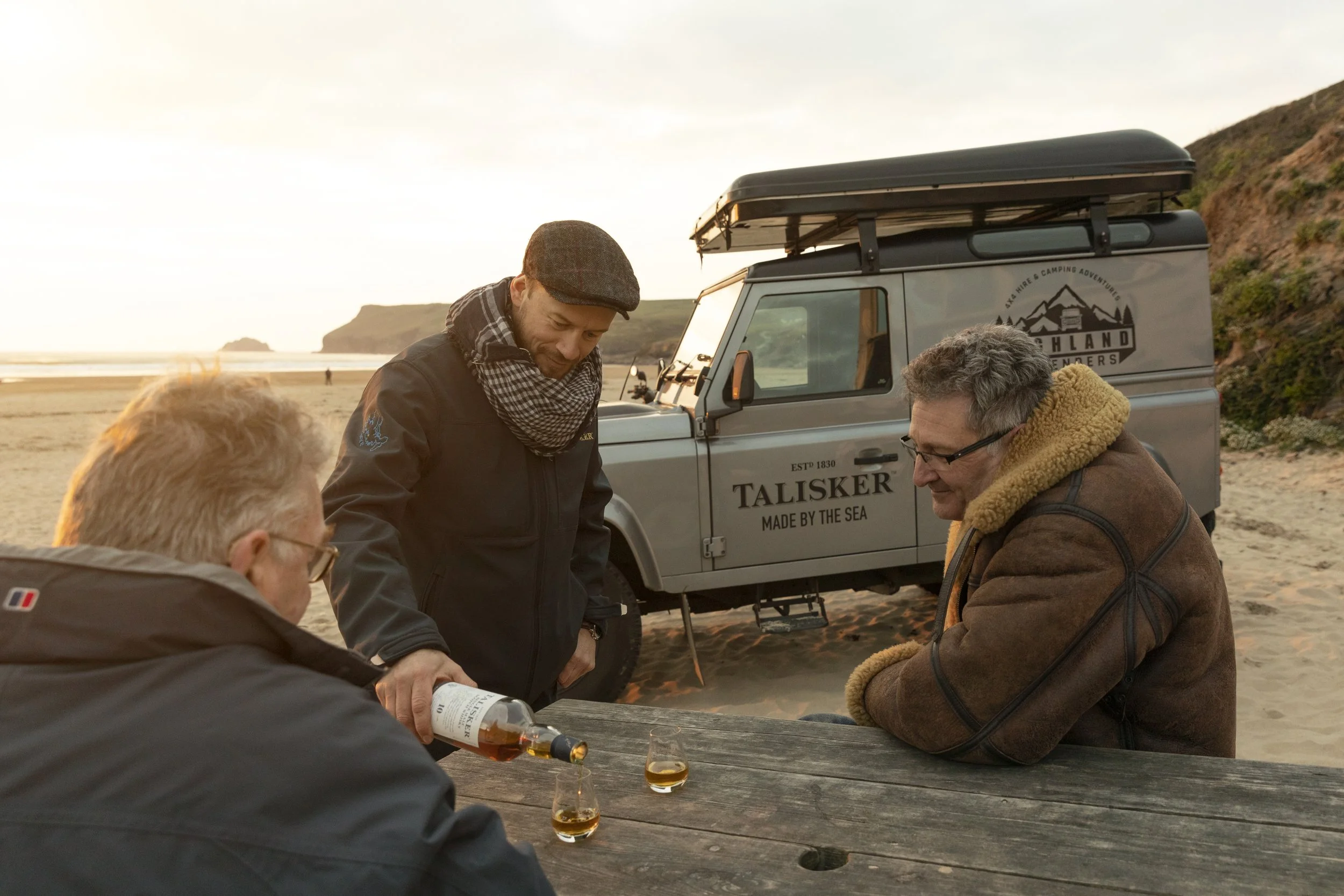 Three men at a beachside outdoor table near a land rover, with one pouring a drink and the others sitting, during sunset.