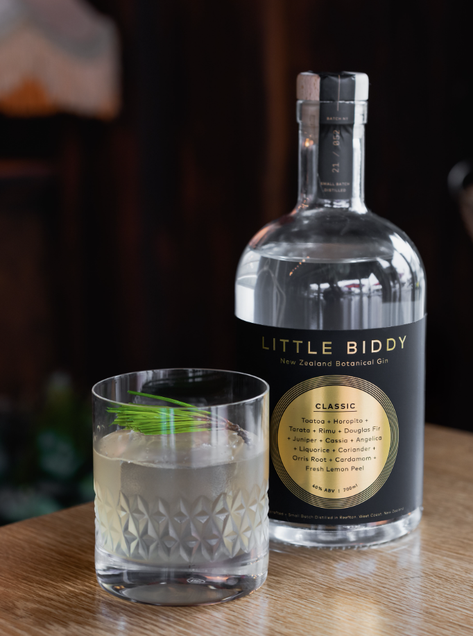 A cocktail in a short glass garnished with a sprig of chive, placed next to a bottle of Little Biddy New Zealand Botanical Gin on a wooden surface, with a dark background.