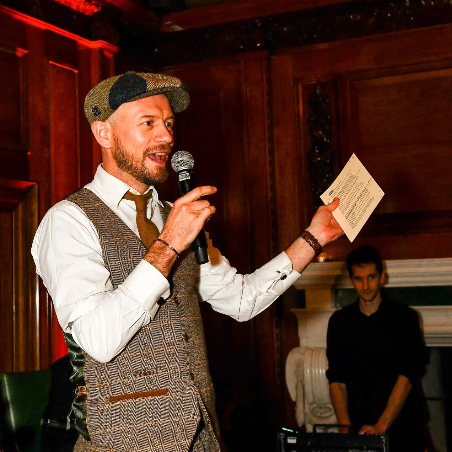 A man speaking into a microphone, wearing a vintage-style cap, dress shirt, tie, and vest. He is holding a piece of paper and appears to be giving a speech or toast at an event in a wood-paneled room. In the background, a woman stands near a fireplac