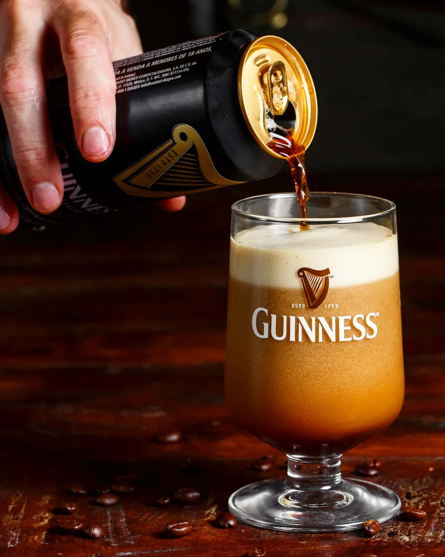 A glass of stout beer branded Guinness on a wooden surface with coffee beans scattered around, being poured from a can.