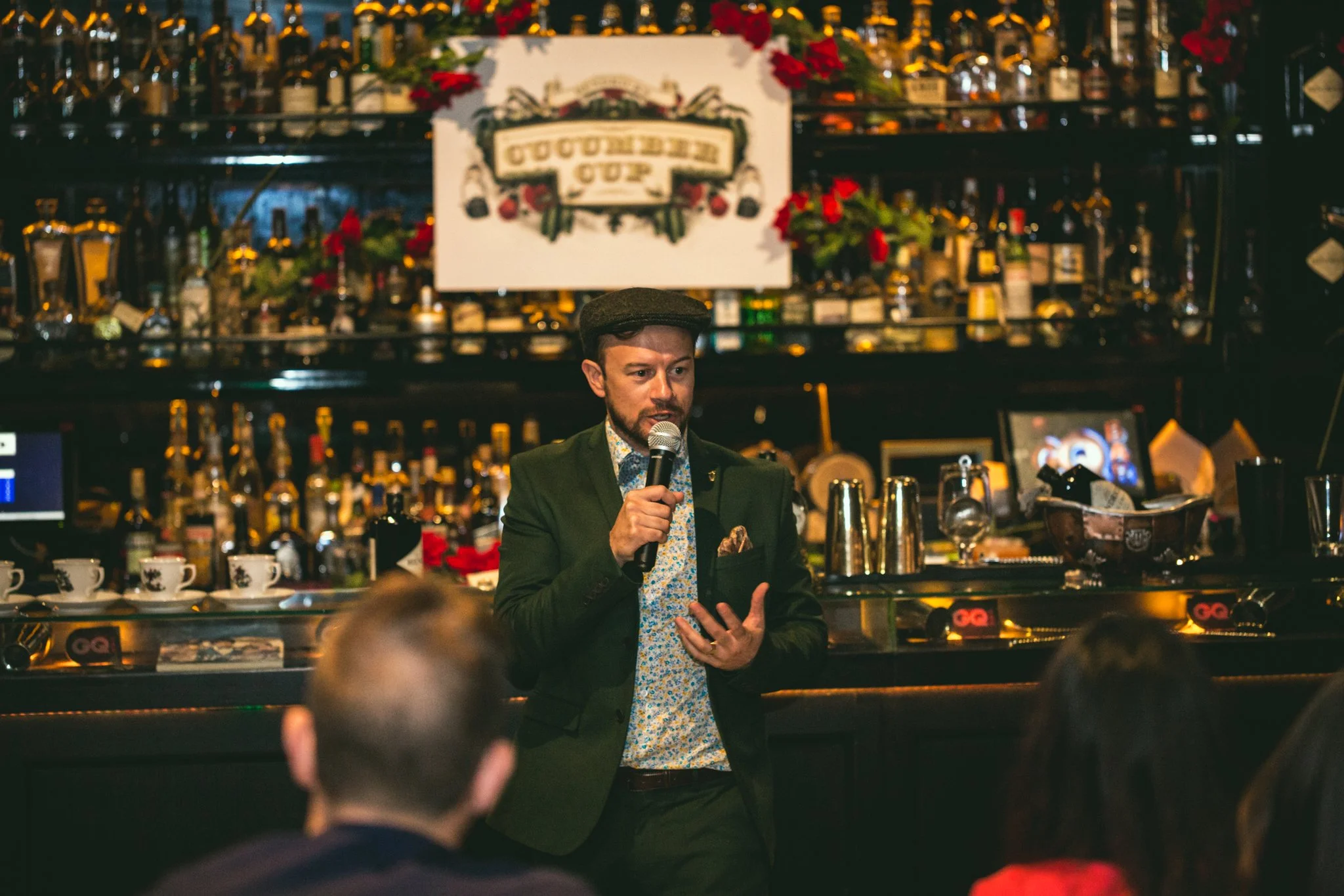 A man wearing a green blazer and floral shirt holding a microphone speaking to an audience at a bar or restaurant. The bar behind him has bottles and glasses, with a sign that reads 'Oktoberfest'. The setting appears to be cozy and warm, possibly during a special event or celebration.