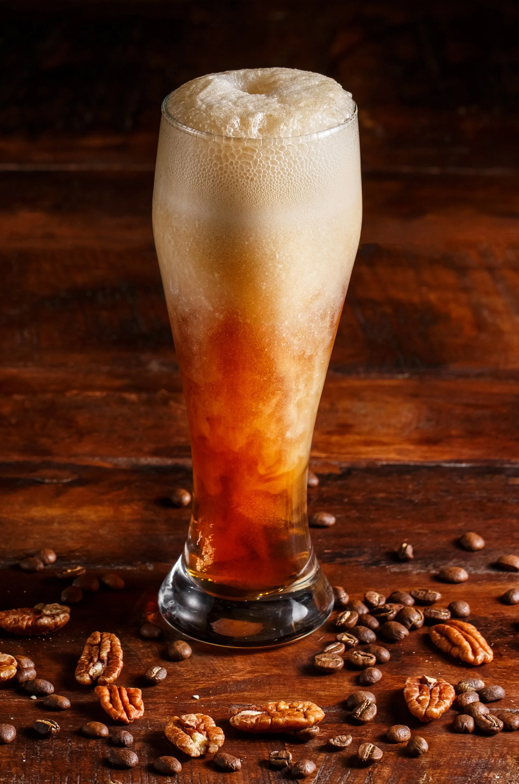 A glass of iced coffee topped with foam, on a wooden surface with scattered coffee beans and pecans.