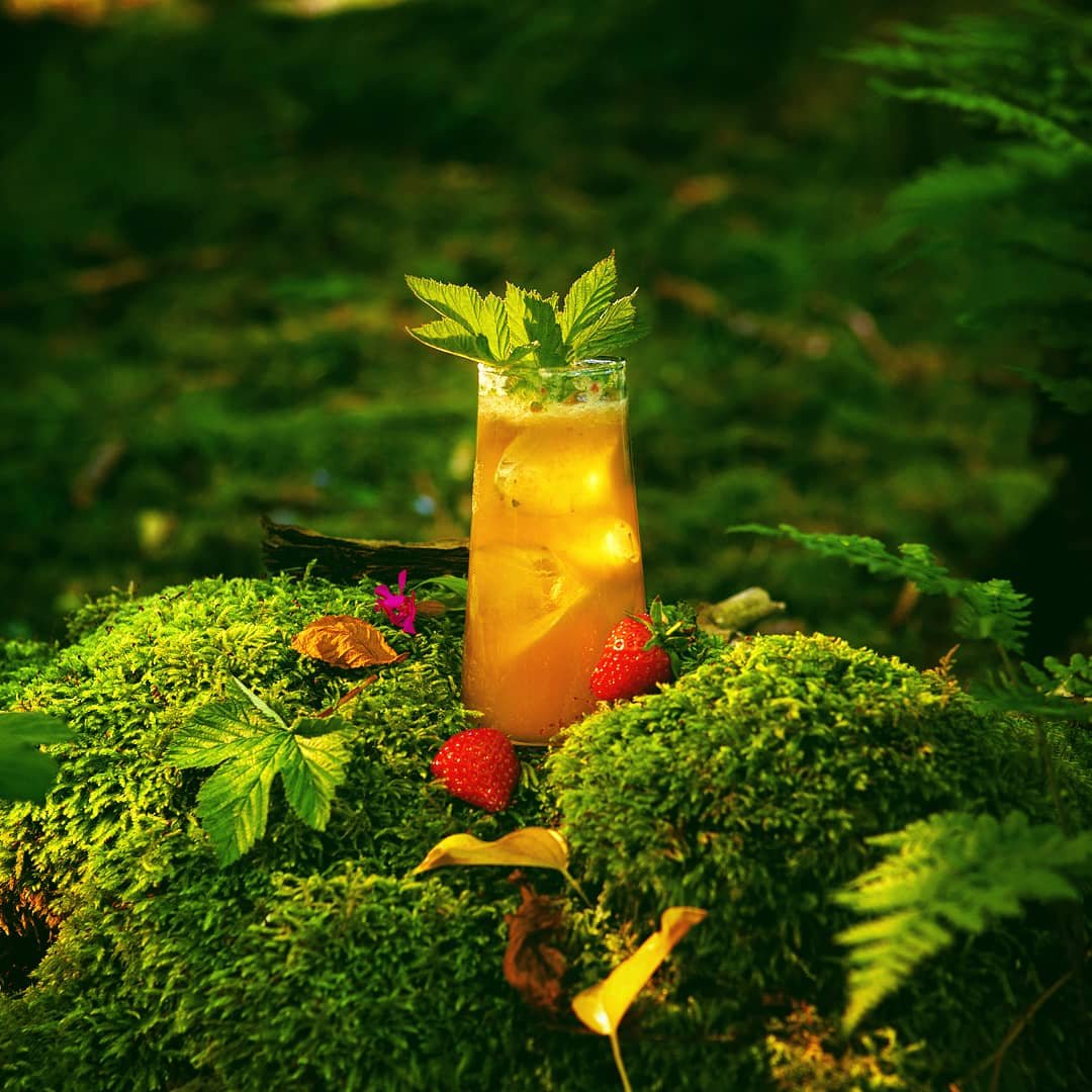 A glass of iced tea with mint leaves on top, surrounded by green moss, strawberries, and fallen leaves in a natural outdoor setting.