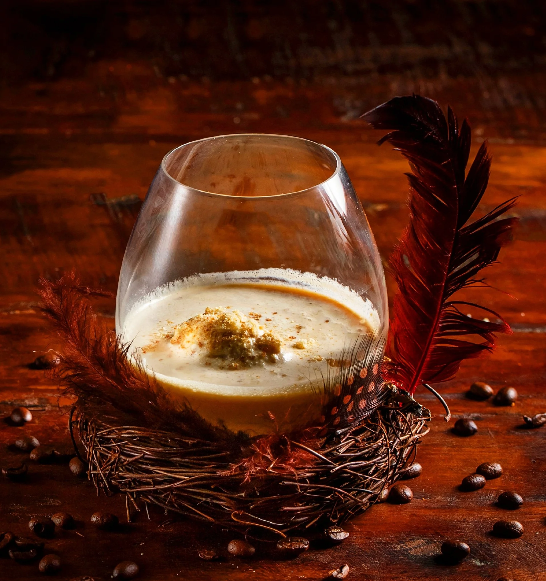 A glass of creamy coffee or latte with foam and a dusting of cinnamon or cocoa powder, nestled in a bird's nest with red and black feathers, surrounded by scattered coffee beans on a wooden surface.