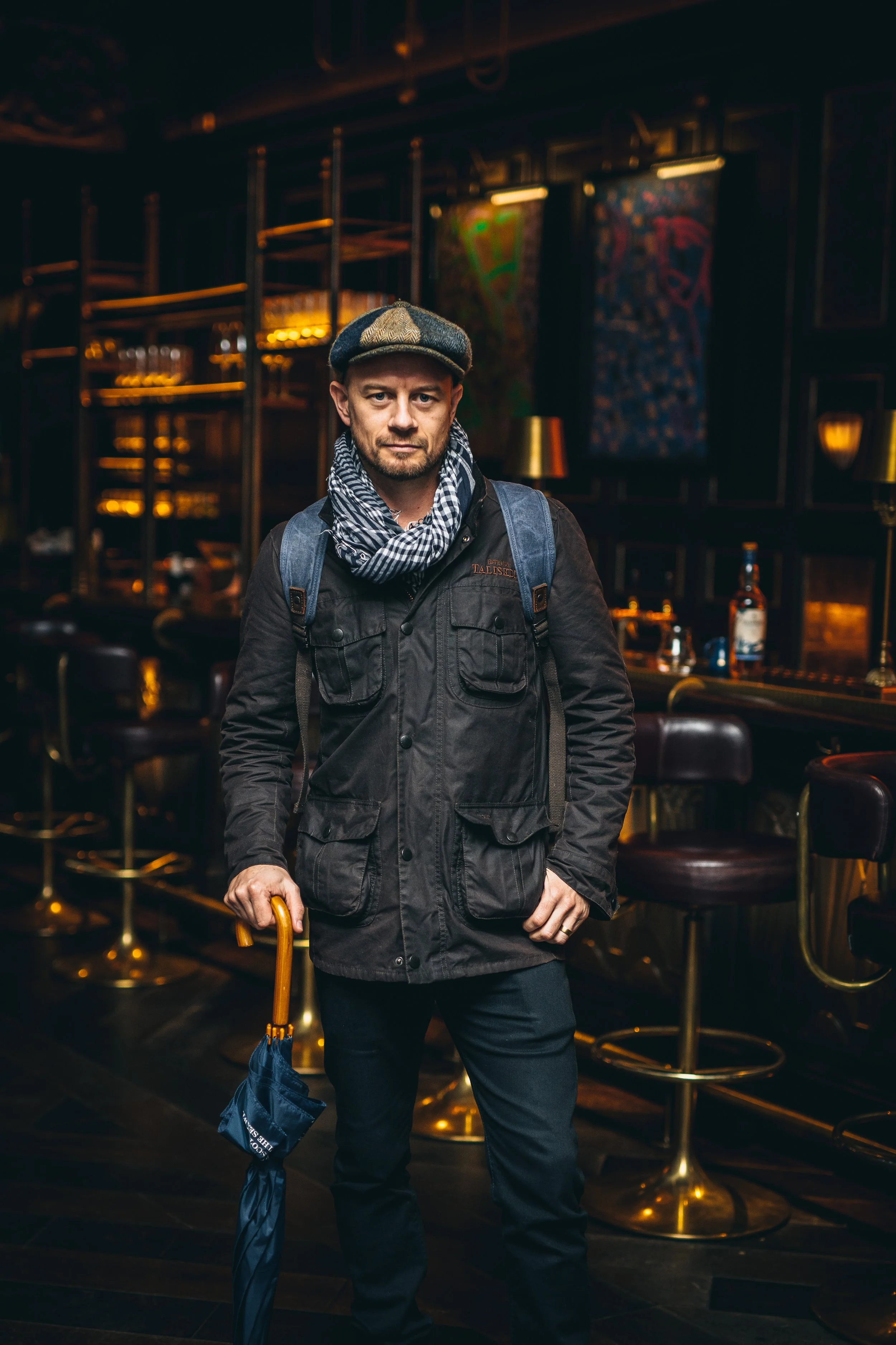 A man stands inside a dimly lit bar, holding a closed umbrella in his right hand. He wears a dark jacket, a gray beret, and a black-and-white checkered scarf. The background features bar stools, bottles, lamps, and colorful abstract art on the walls.