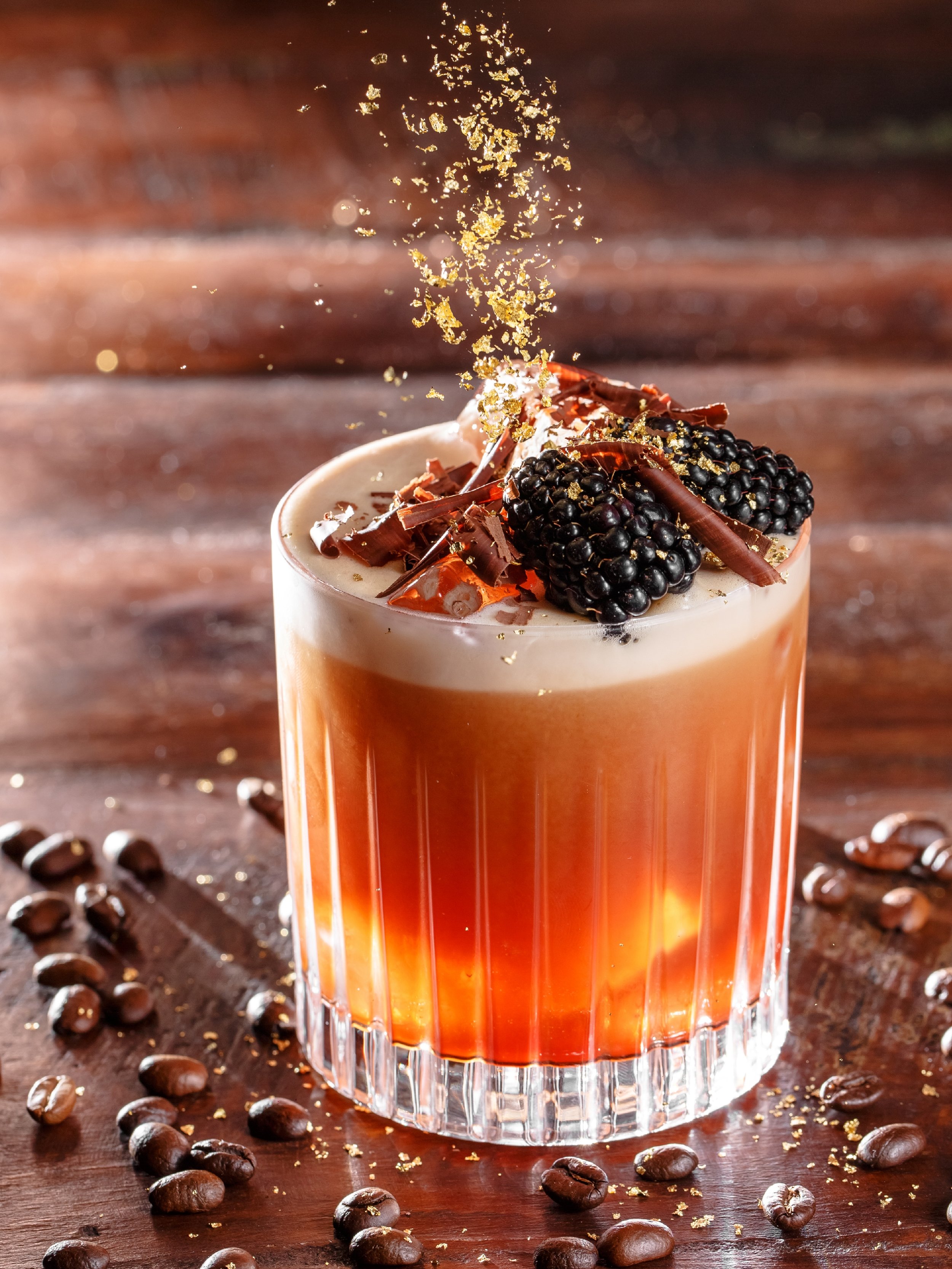 A glass of coffee drink topped with blackberries, chocolate shavings, and gold leaf, with coffee beans scattered on a wooden surface.