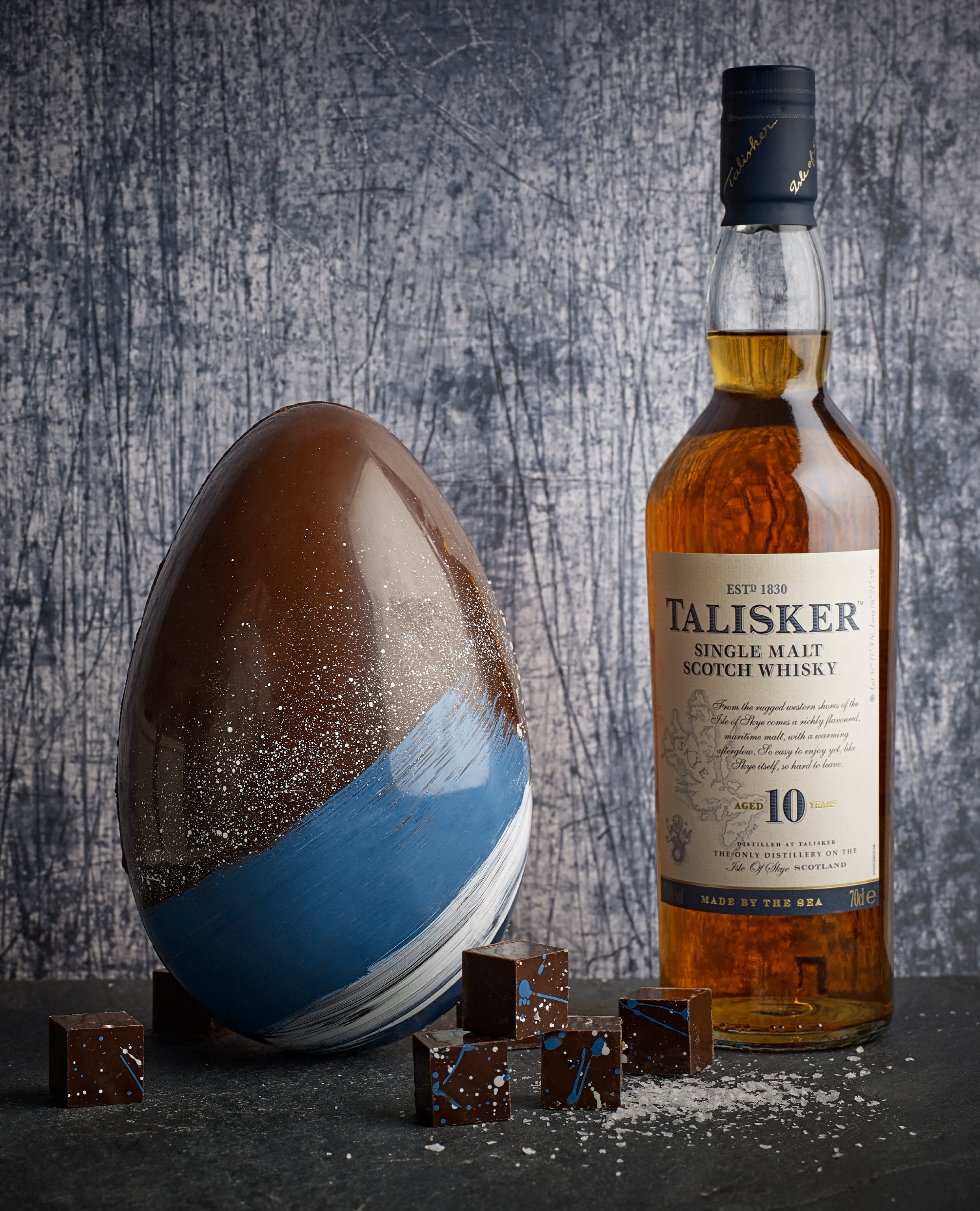 Decorative chocolate egg with blue and white paint splatter, a bottle of Talisker single malt scotch whisky, and several small chocolate pieces with blue and white splatters on a dark surface against a textured gray background.