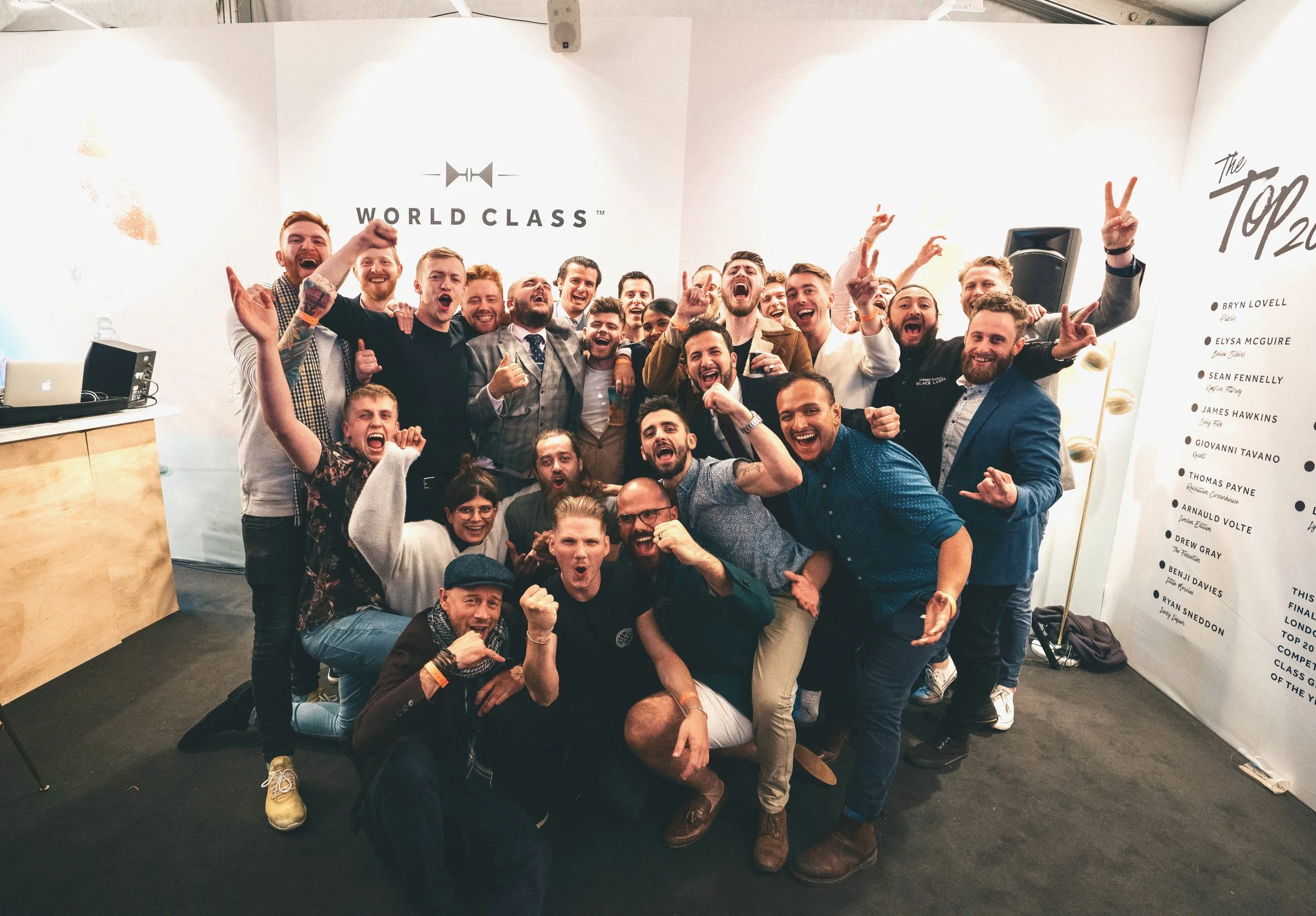 A group of cheerful people posing together indoors, celebrating with many smiling faces and raised fists and hands, with a backdrop that reads 'WORLD CLASS' and a list of names.