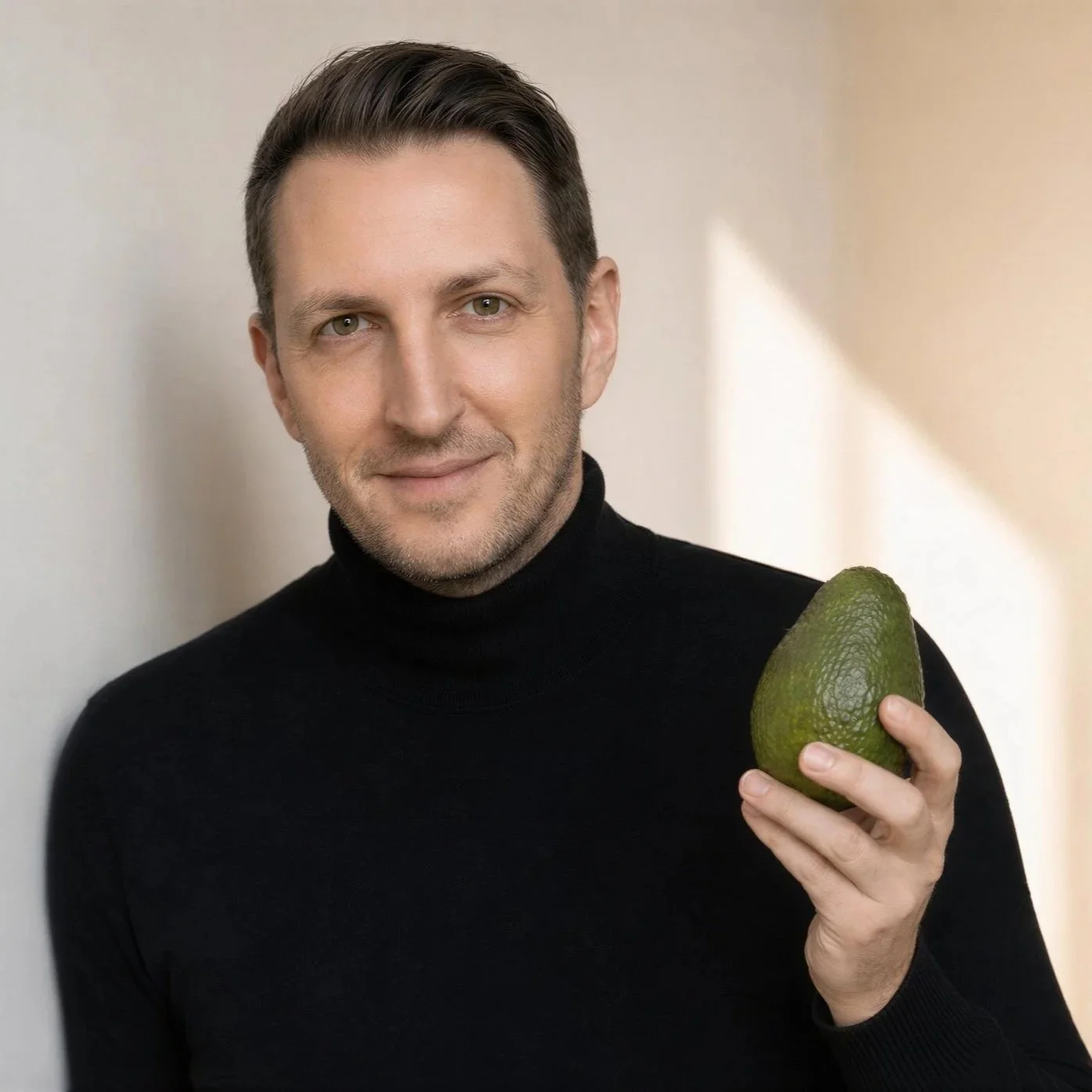 Ein Mann mit braunem Haar und Bart trägt einen schwarzen Rollkragenpullover und hält eine Avocado in der Hand. Er lächelt leicht und steht vor einem hellen, schlichten Hintergrund.