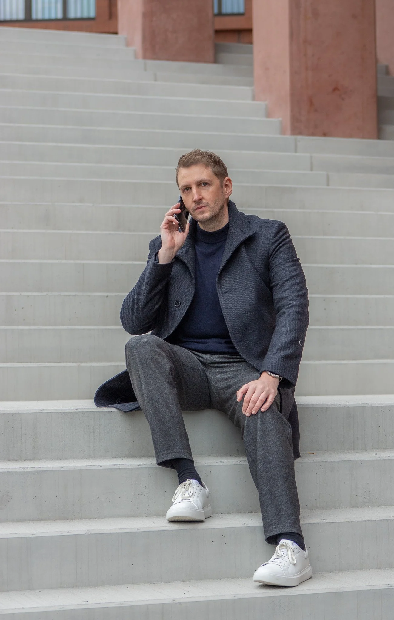 Mann sitzt auf Treppen, spricht am Telefon, trägt dunkle Jacke, schwarze Hemd, graue Hosen, weiße Sneakers, Blick nach oben.