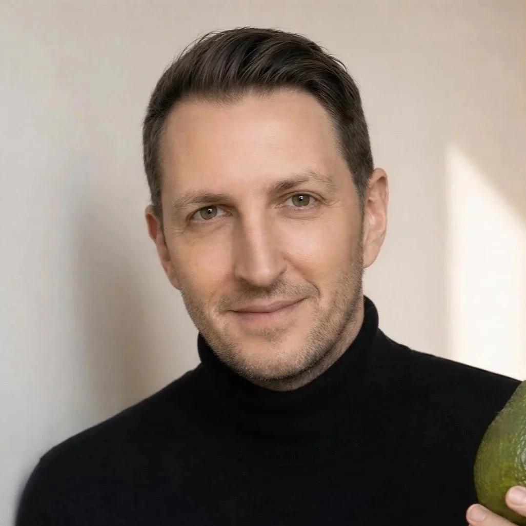 Porträt eines jungen Mannes mit dunklem Turtleneck und dunklem Jackett, lächelnd, vor hellem Hintergrund.