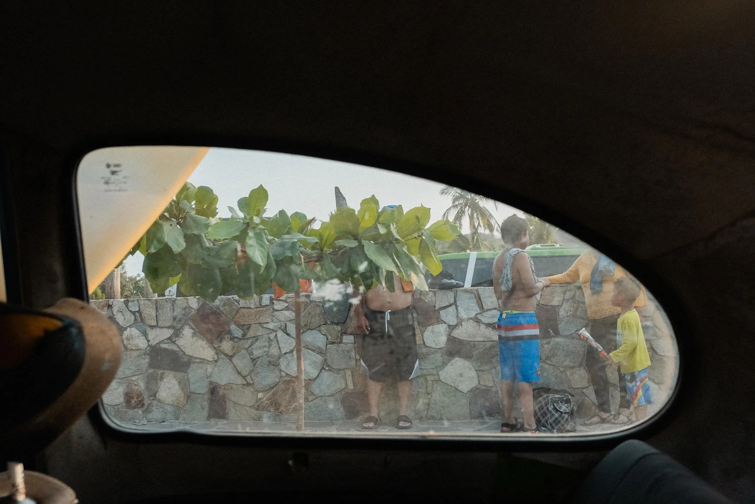 Photograph through the rear passenger window of a Volkswagen Beetle, showing several tourists in swimwear in Mexico, part of Irving Villegas’ LÄUFT photography project.