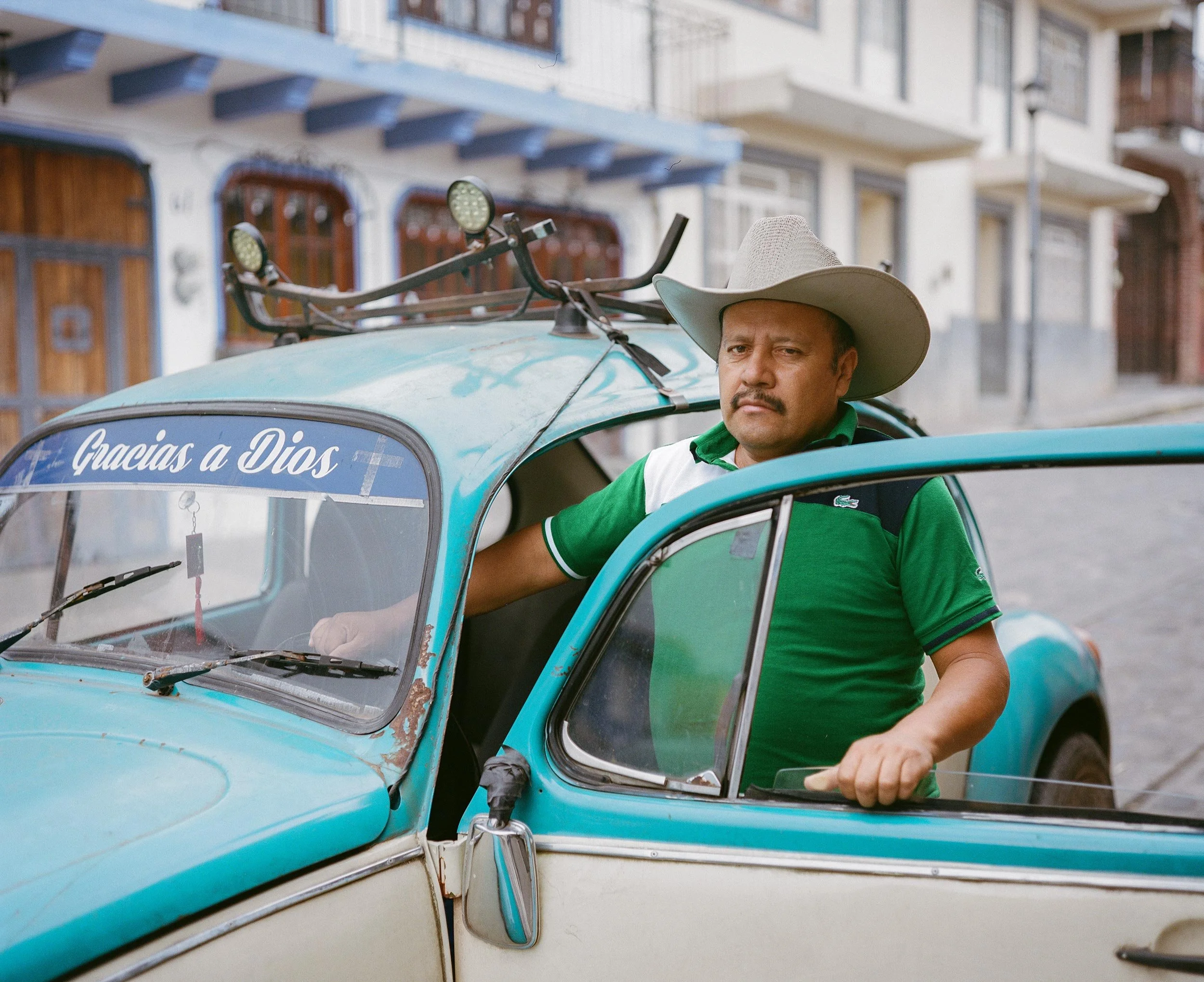 Portrait of a man sitting in a turquoise Volkswagen Beetle in Mexico, with “Gracias a Dios” written on the front of the car, photographed by Irving Villegas for his documentary project LÄUFT.