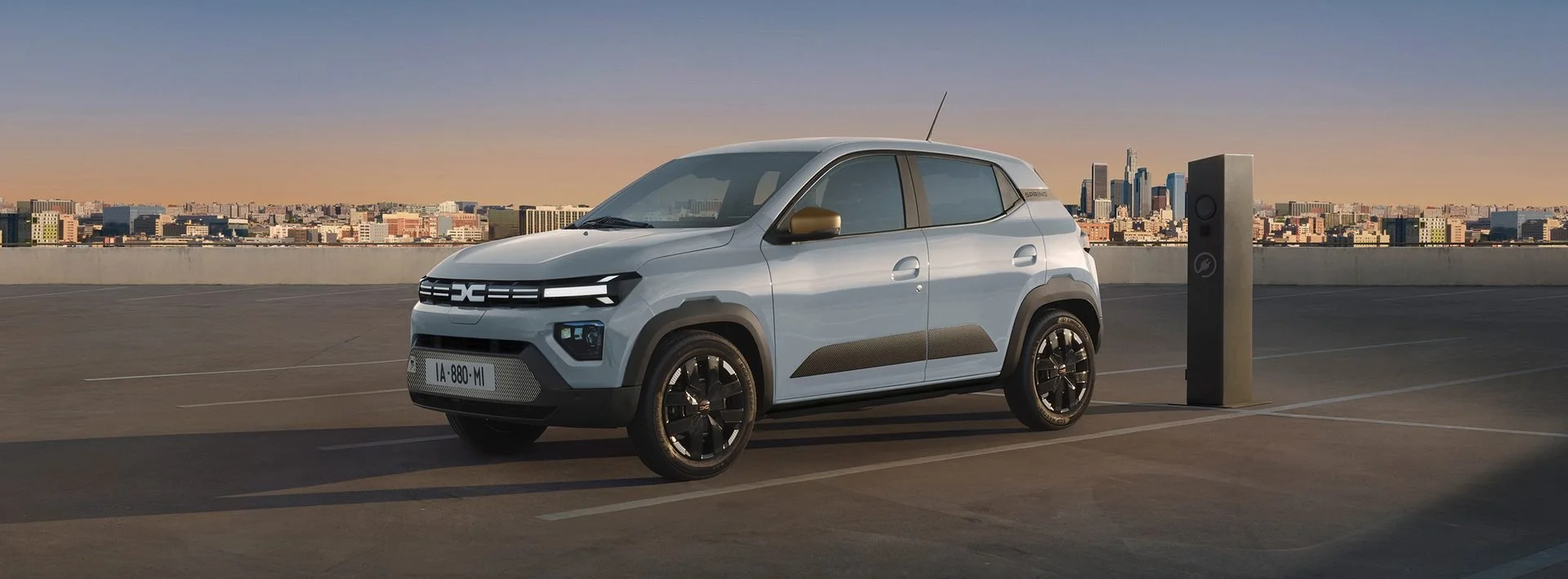 Dacia Spring cinzento estacionado junto a um posto de carregamento, com a cidade ao fundo ao pôr do sol