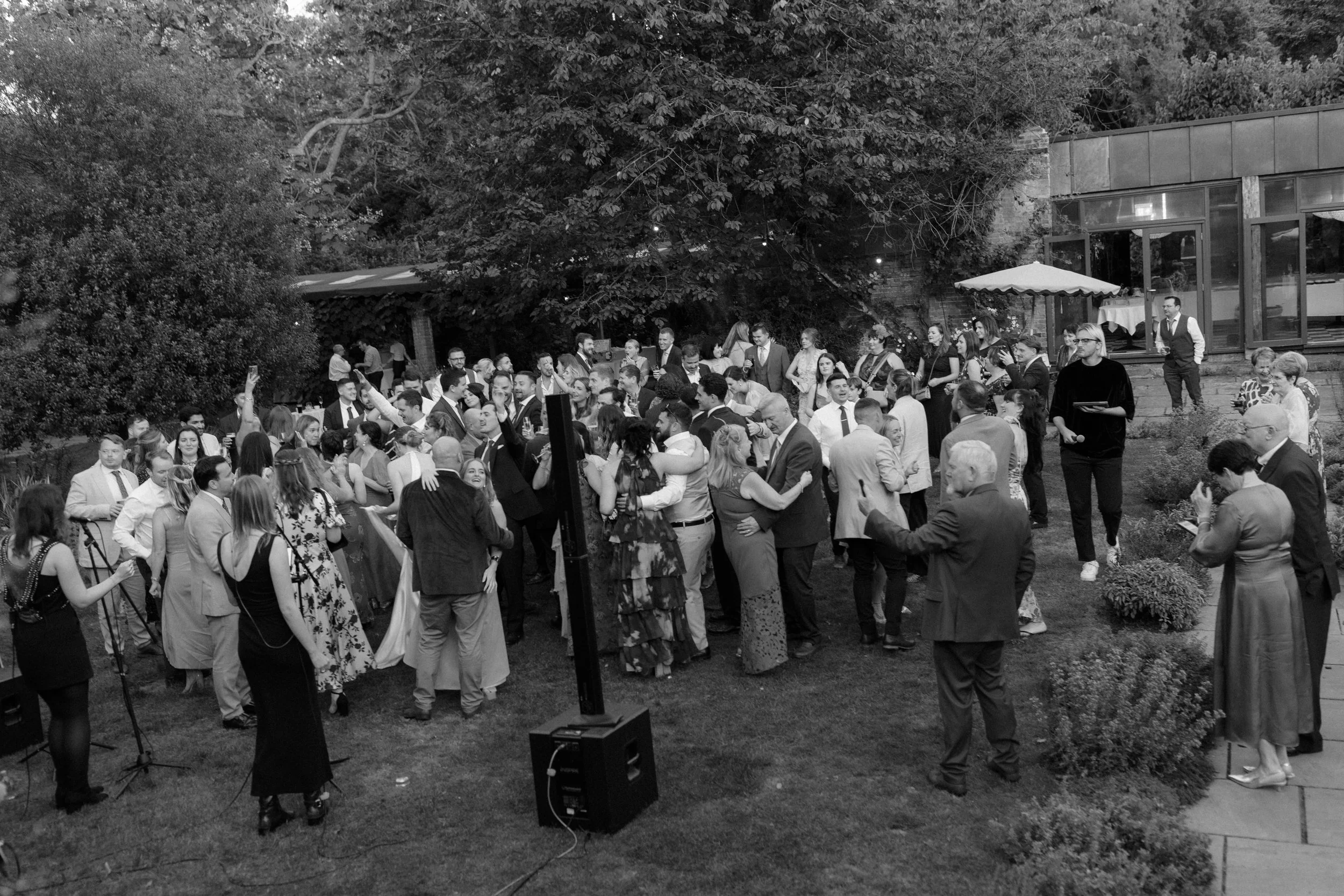 A large outdoor party or wedding reception with many people dancing, talking, and taking photos on a grassy area surrounded by trees and bushes. Some guests are hugging, and there is a person holding a microphone and a speaker in the foreground.
