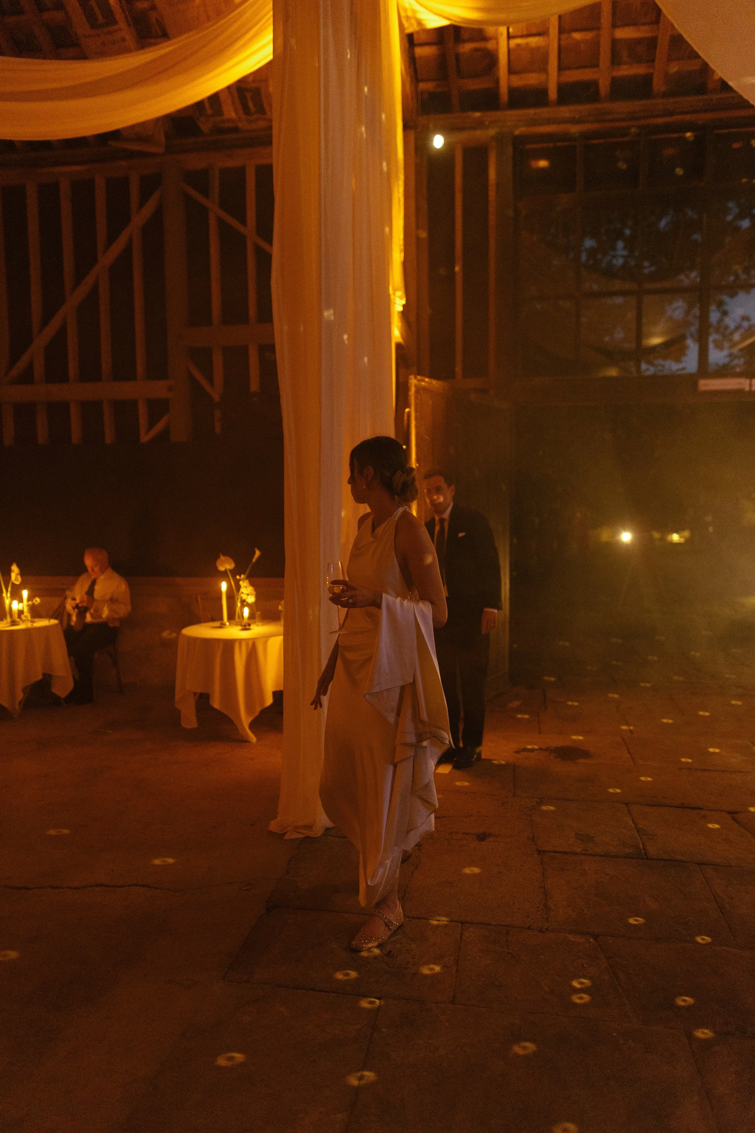 A woman in an elegant dress holding a glass at a dimly lit, rustic indoor event with tables set with flowers and candles.