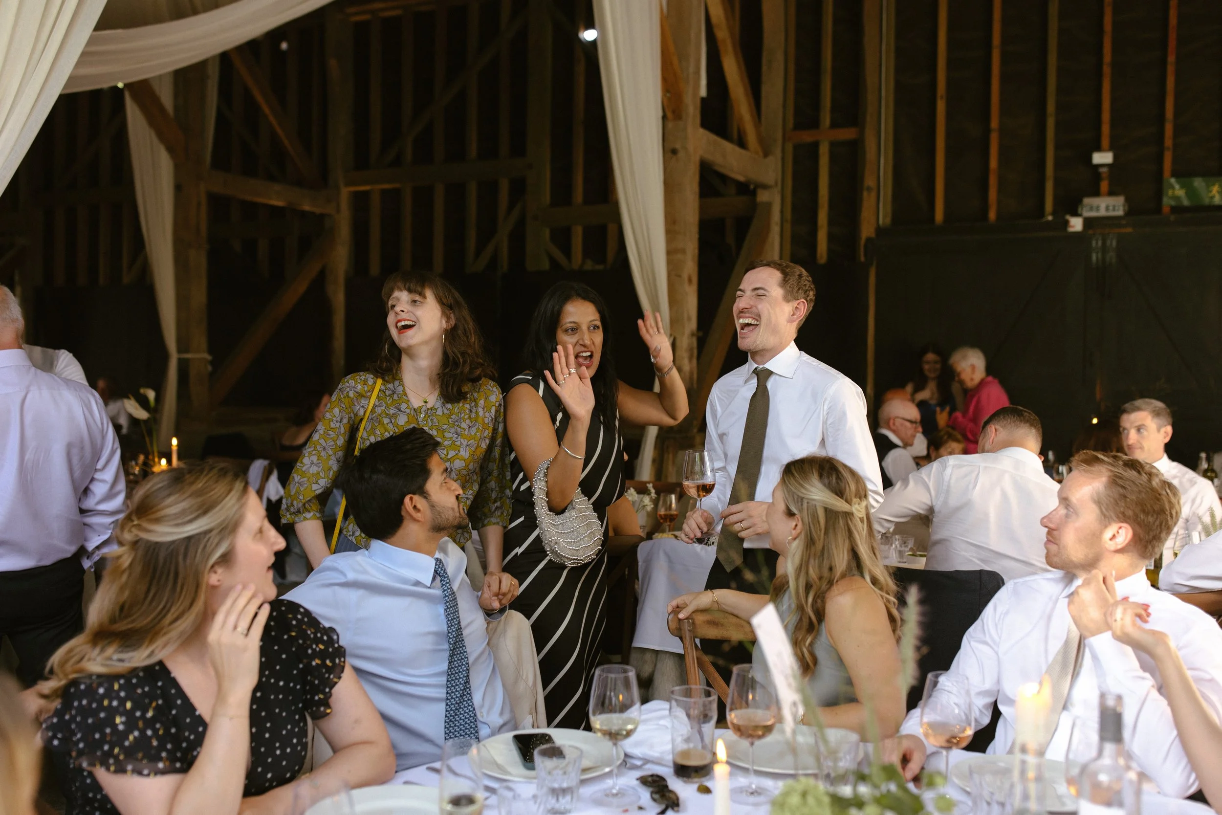 Group of people at a wedding reception, some standing and some seated, laughing and chatting in a rustic barn setting.