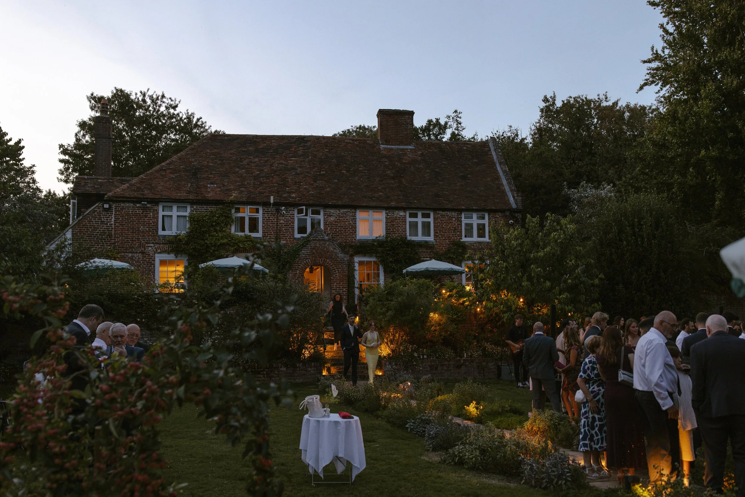 Outdoor evening gathering at a garden party with guests socializing near a brick house, illuminated by warm outdoor lighting, with trees and a sky in the background.
