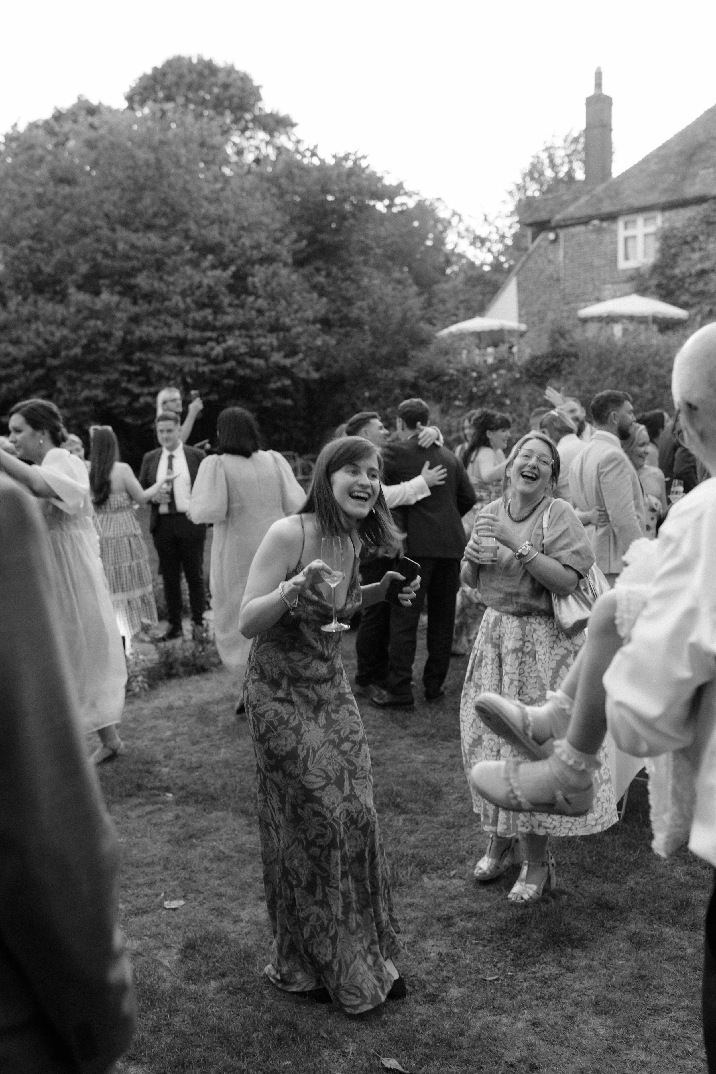 People at an outdoor social gathering, smiling and laughing, with some holding drinks. The setting appears to be in a garden or yard with trees and a house in the background.