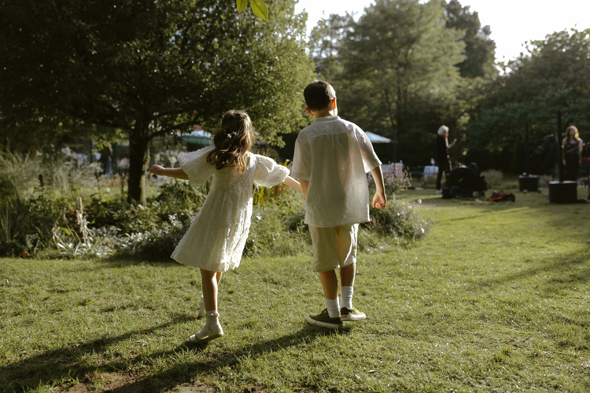 Two children, a girl in a white dress and a boy in matching shorts and shirt, holding hands and walking on a grassy area in a park with trees, with a band or musicians performing in the background.
