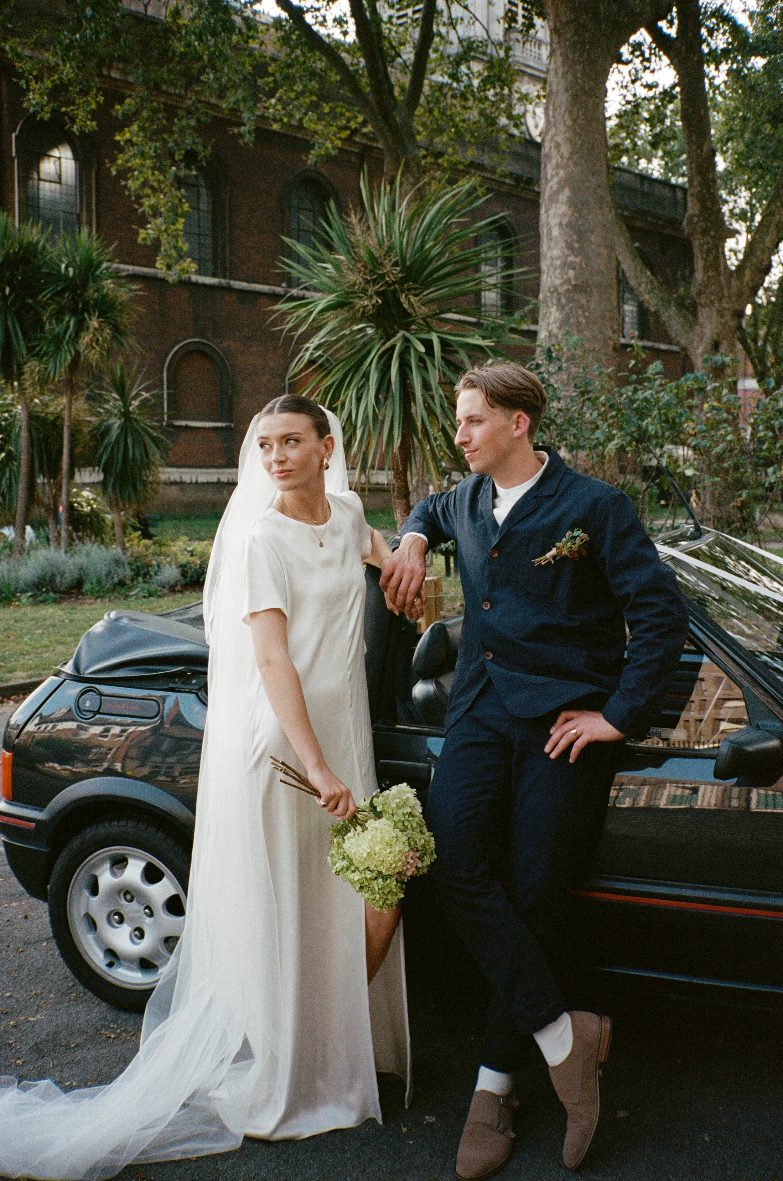 Bride and groom standing next to a black convertible car outdoors, with trees and a Hackney church in the background.