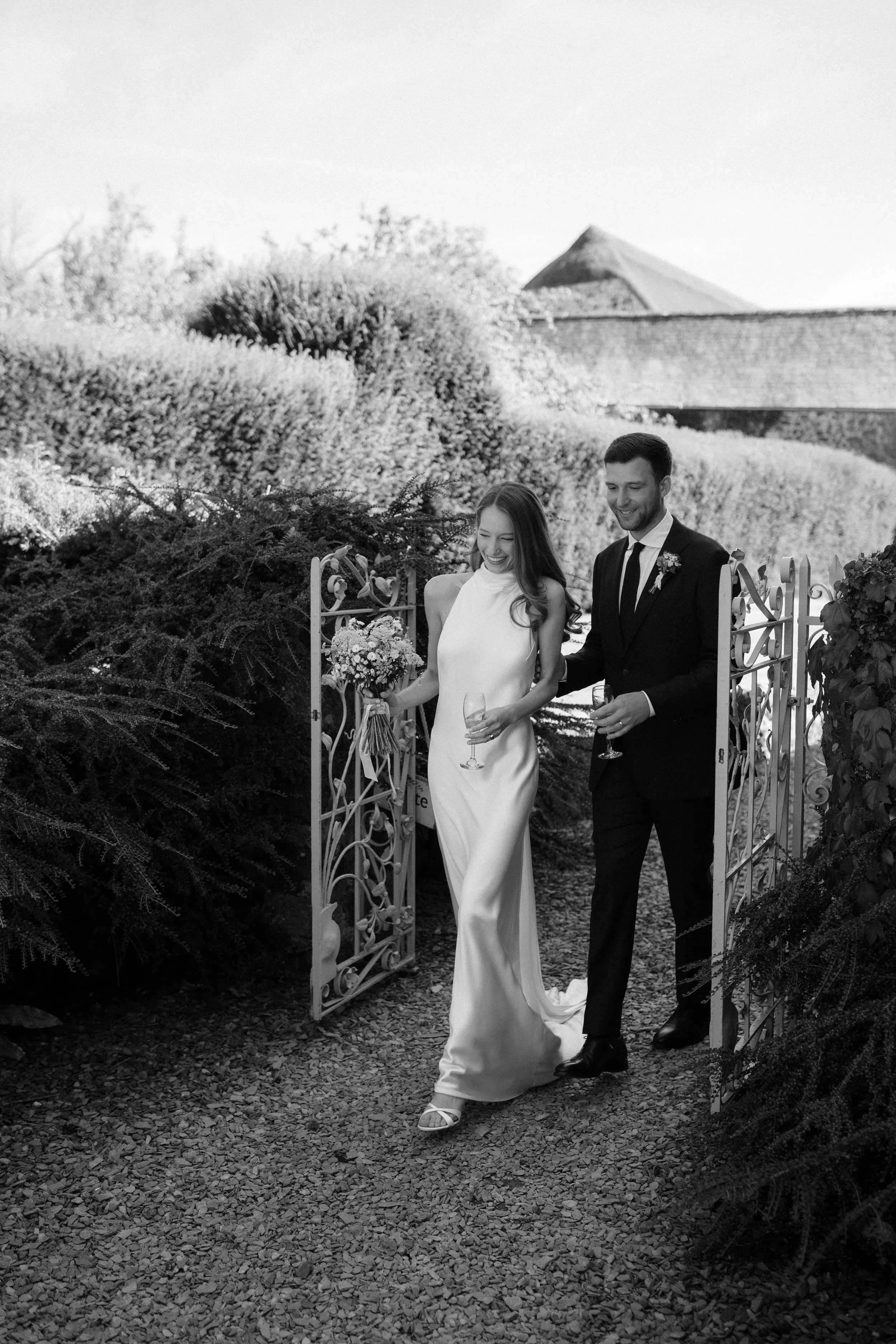 A bride and groom walking out of a gate, smiling and holding glasses, at their wedding outdoors.