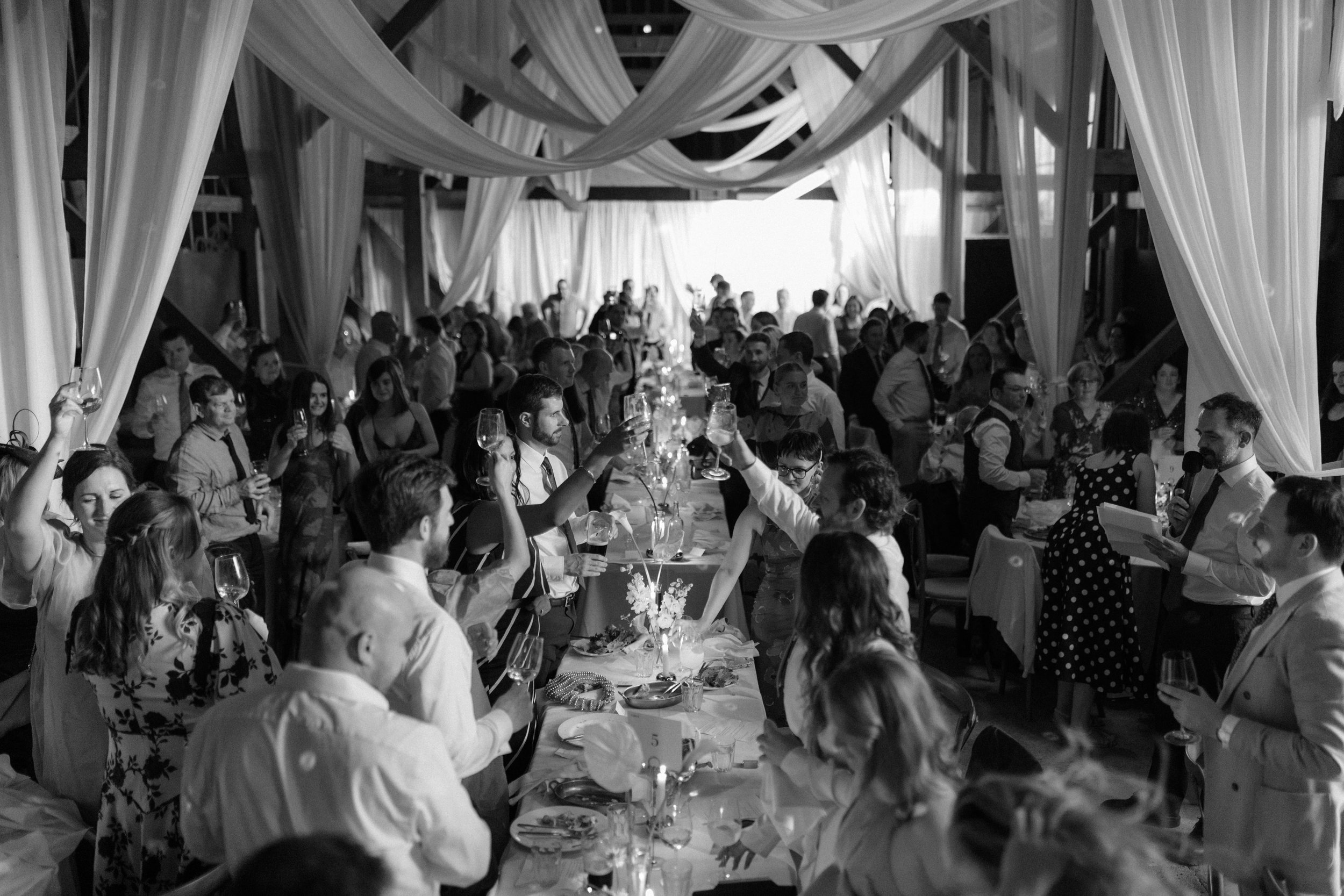 A black and white photo of a lively wedding reception with guests raising glasses in a toast around a long banquet table, decorated with floral centerpieces and surrounded by draped curtains and a large gathering of people.