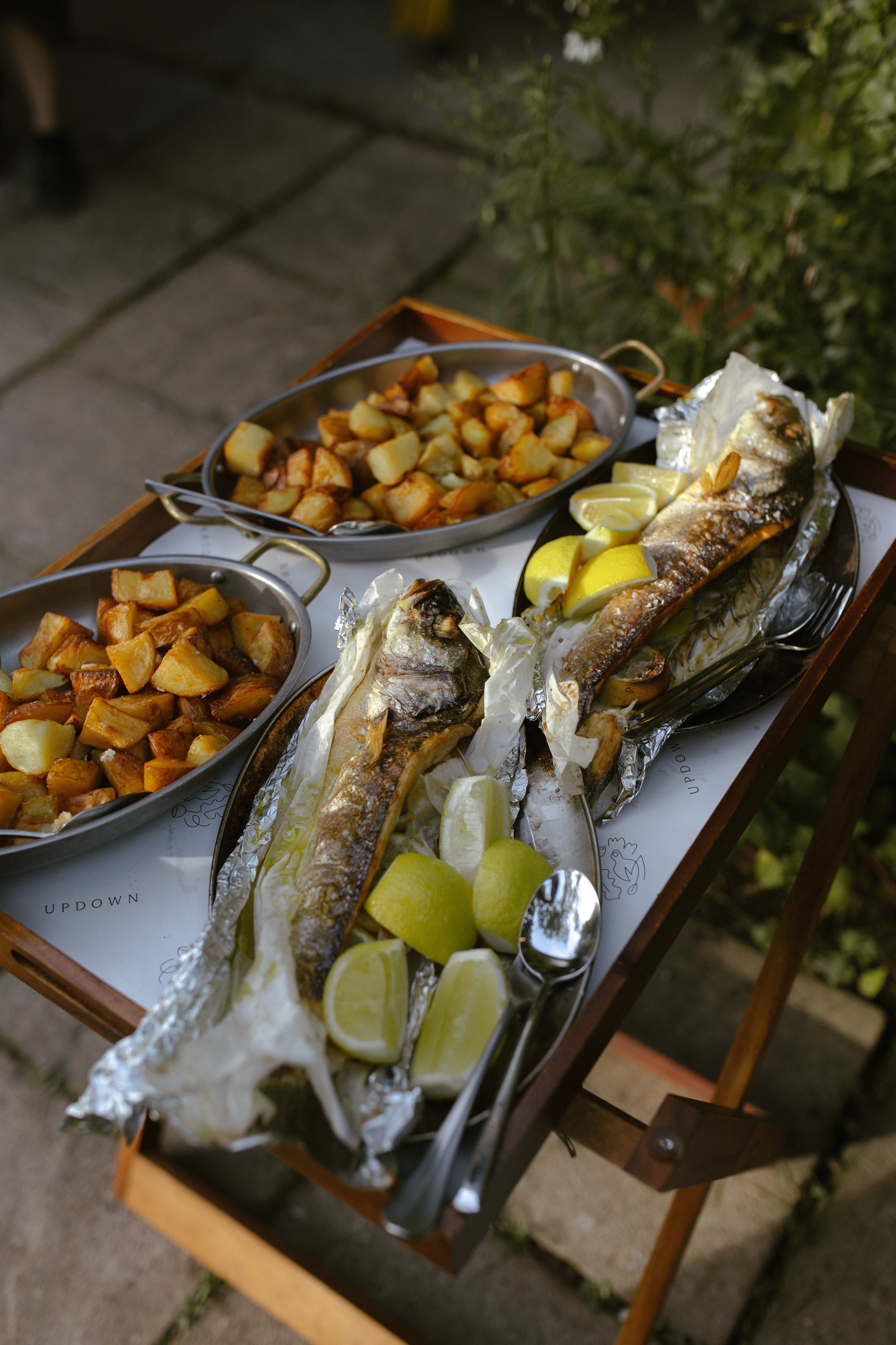 Two grilled fish served with lemon wedges and foil, with roasted potatoes in bowls, on a wooden tray.
