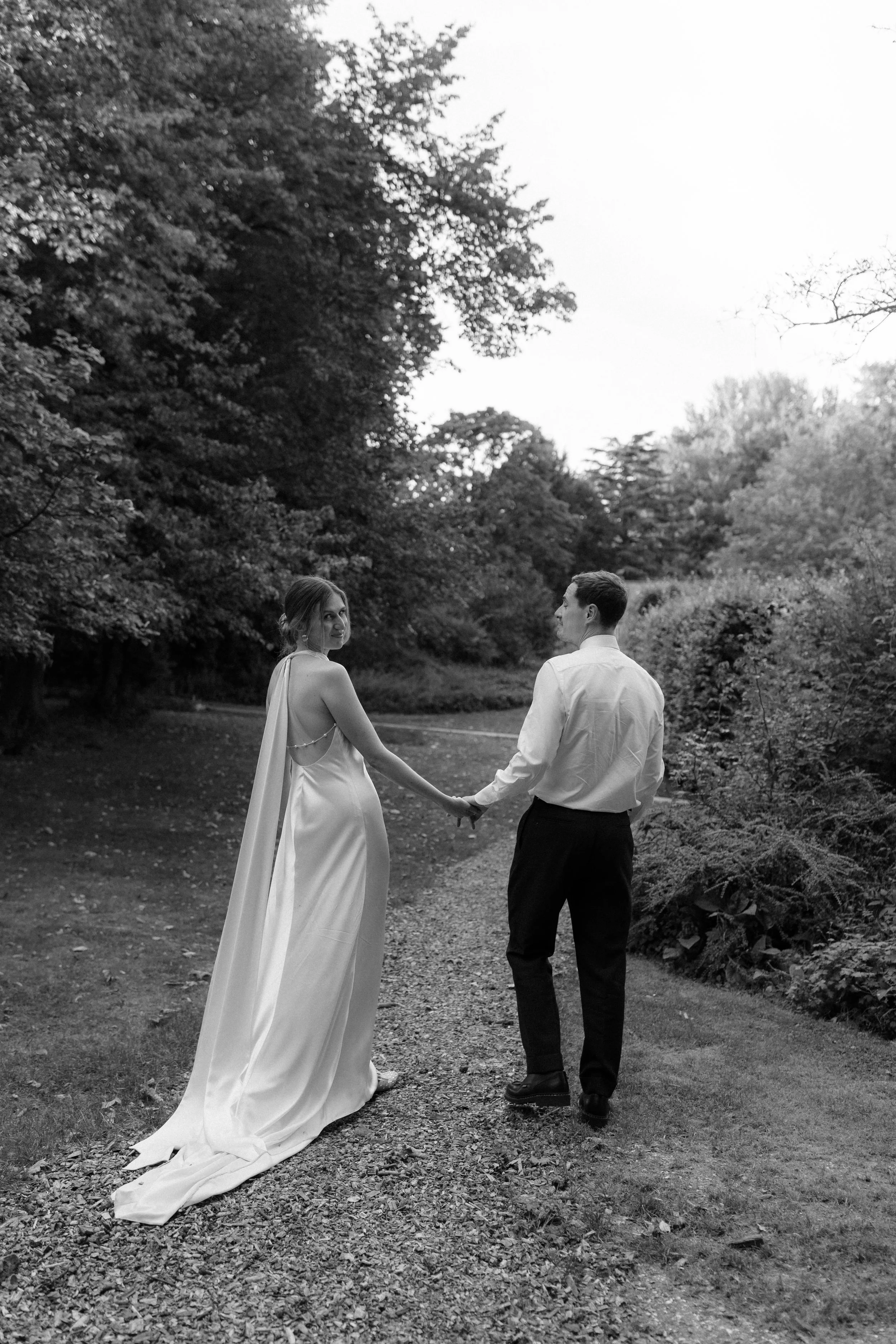 A bride and groom holding hands and walking on a path in a wooded outdoor area, seen from behind in black and white.