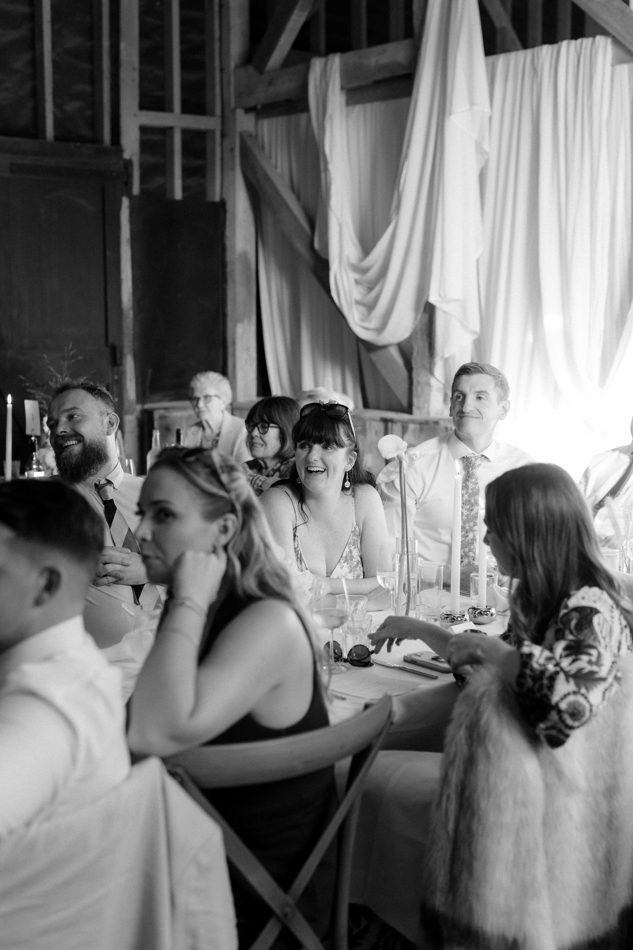 People sitting at a dining table during a celebration, with a woman smiling and others smiling and talking, in a rustic setting with wooden walls and draped fabric.