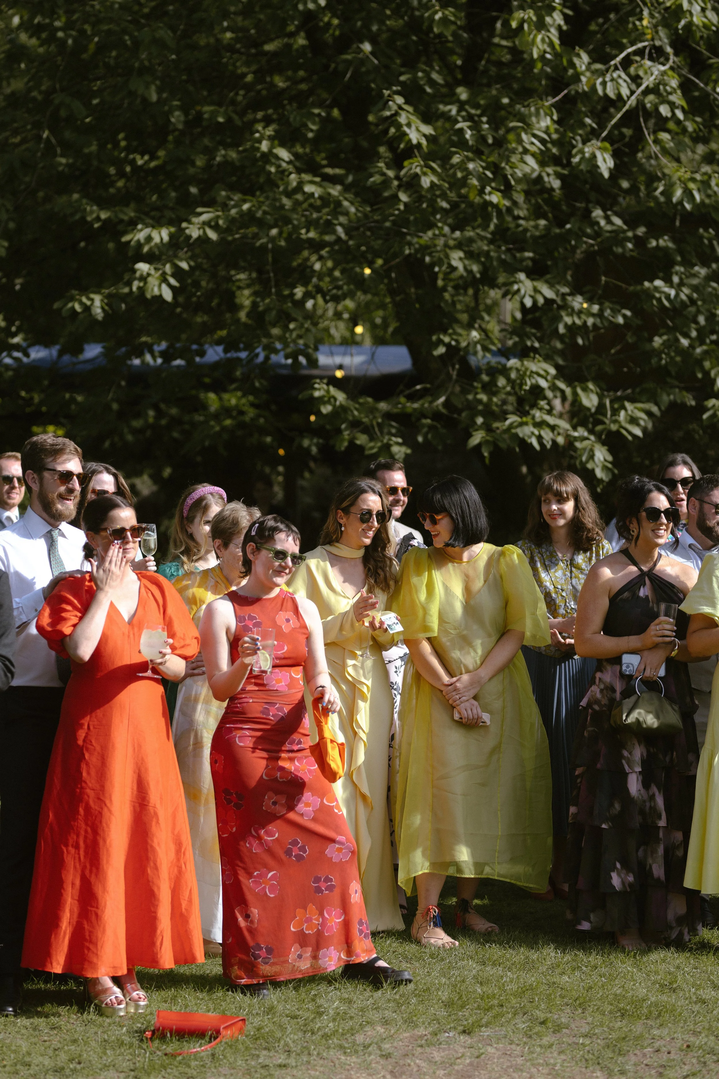 Group of people gathered outdoors for a celebration, dressed in colorful summer outfits, standing on grass under a large tree.