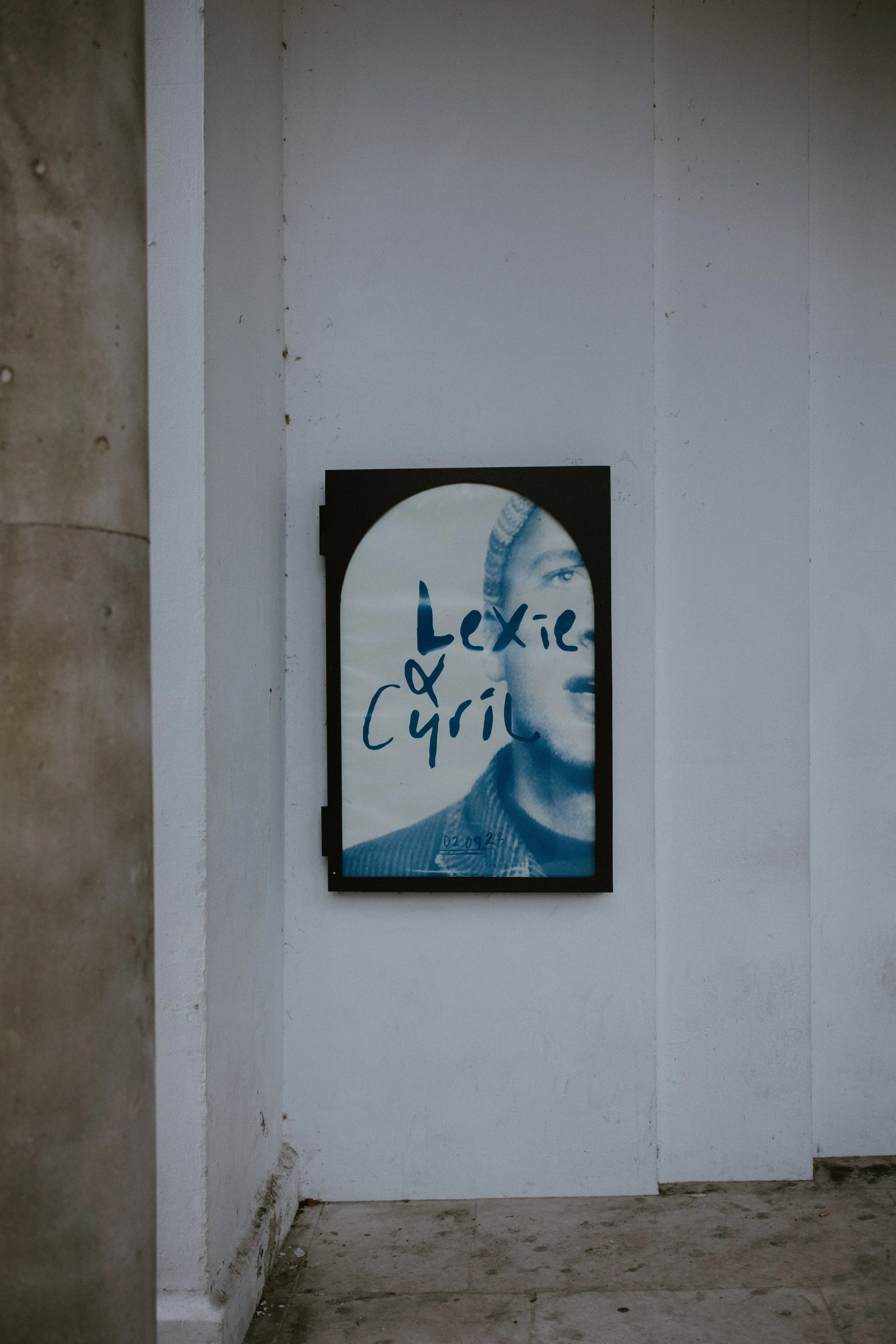 Photo of a framed poster on a white wall in a gallery or exhibit space. The poster features a black and white portrait of a woman with some text written across her face in blue, which reads 'Lexie Girl.'