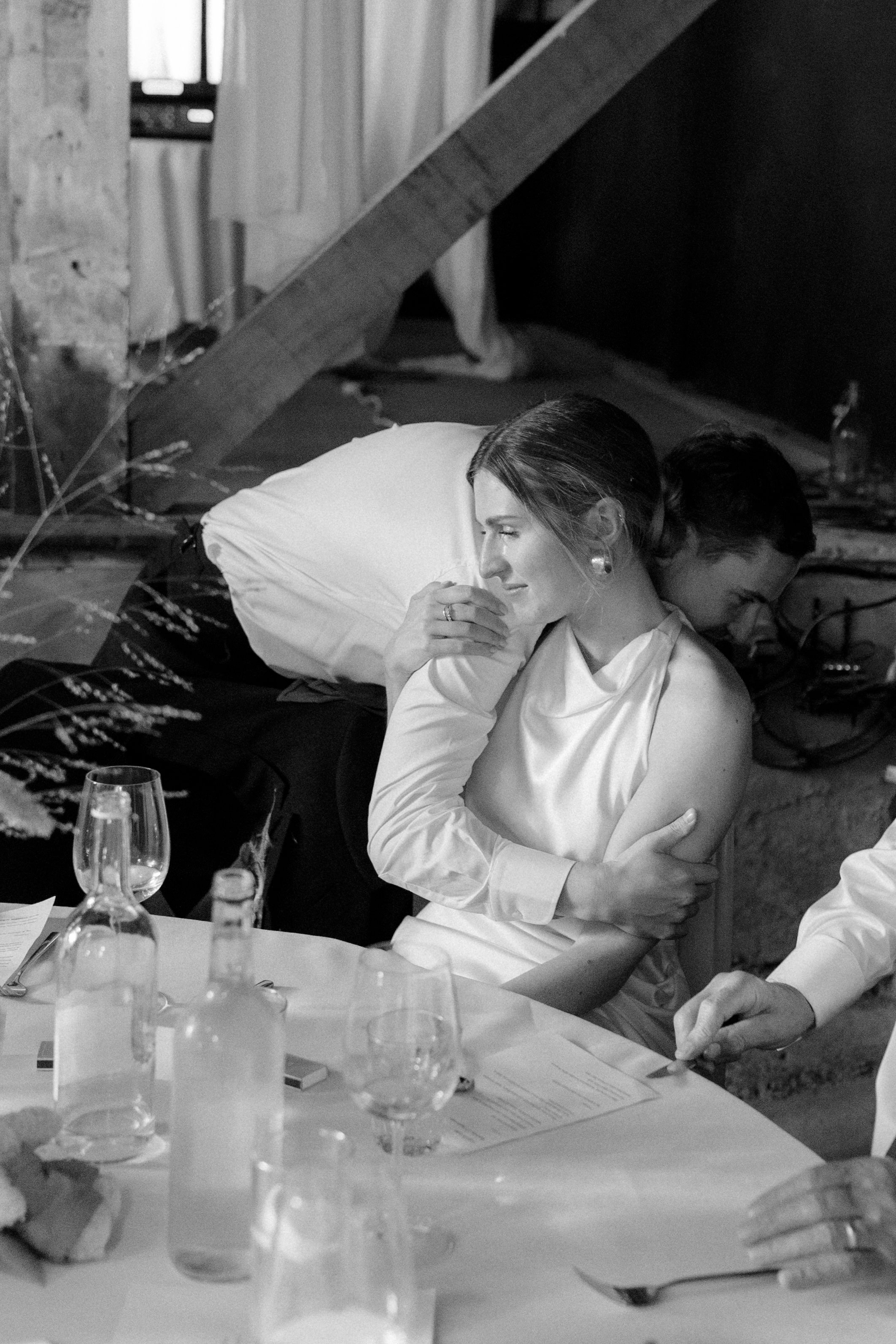 A woman in a sleeveless dress sitting at a table during a formal event, with a man leaning over her shoulder in the background. The table has glasses, bottles, and papers, and the setting appears to be indoors.