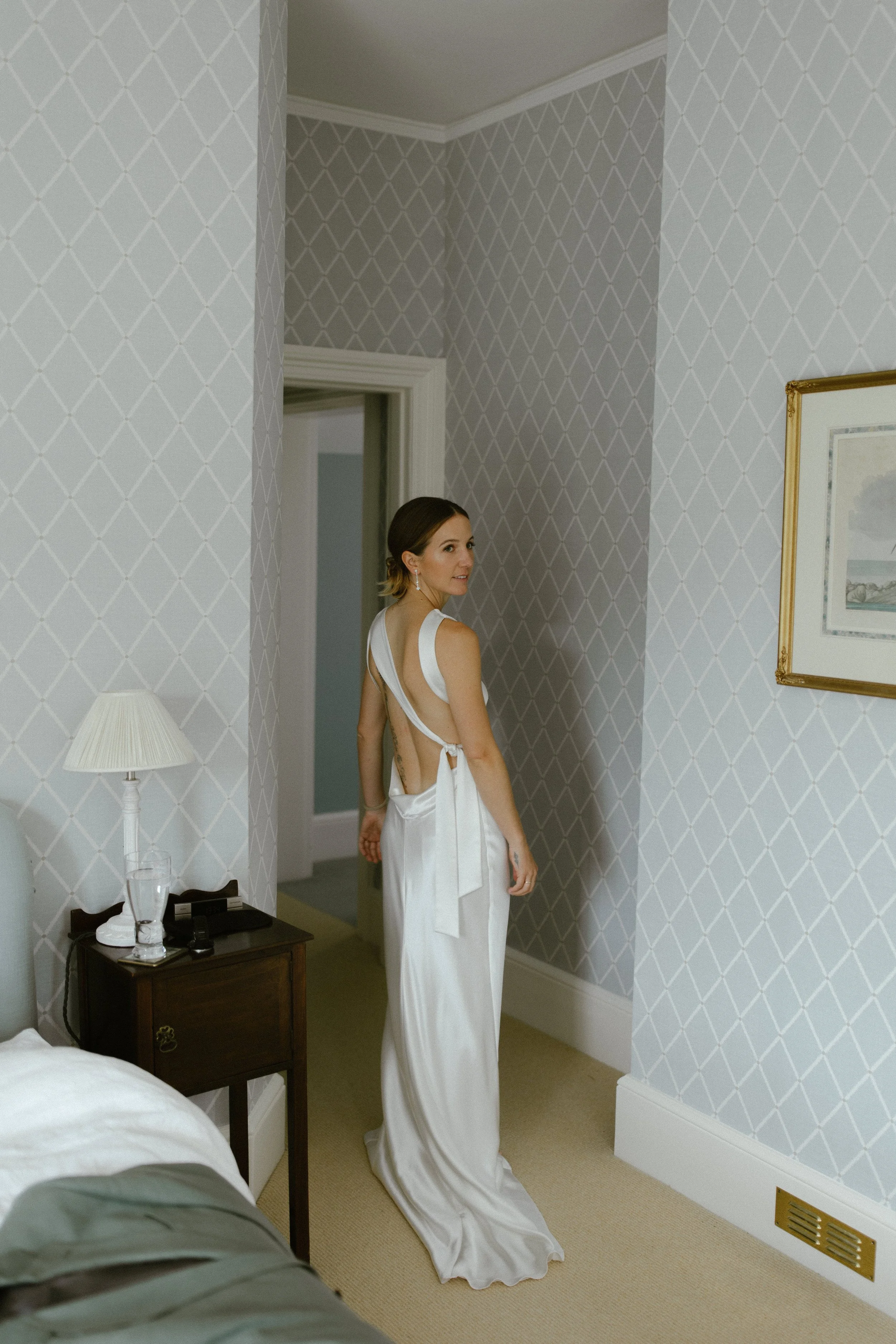 A woman in a white dress standing in a bedroom, looking back over her shoulder.