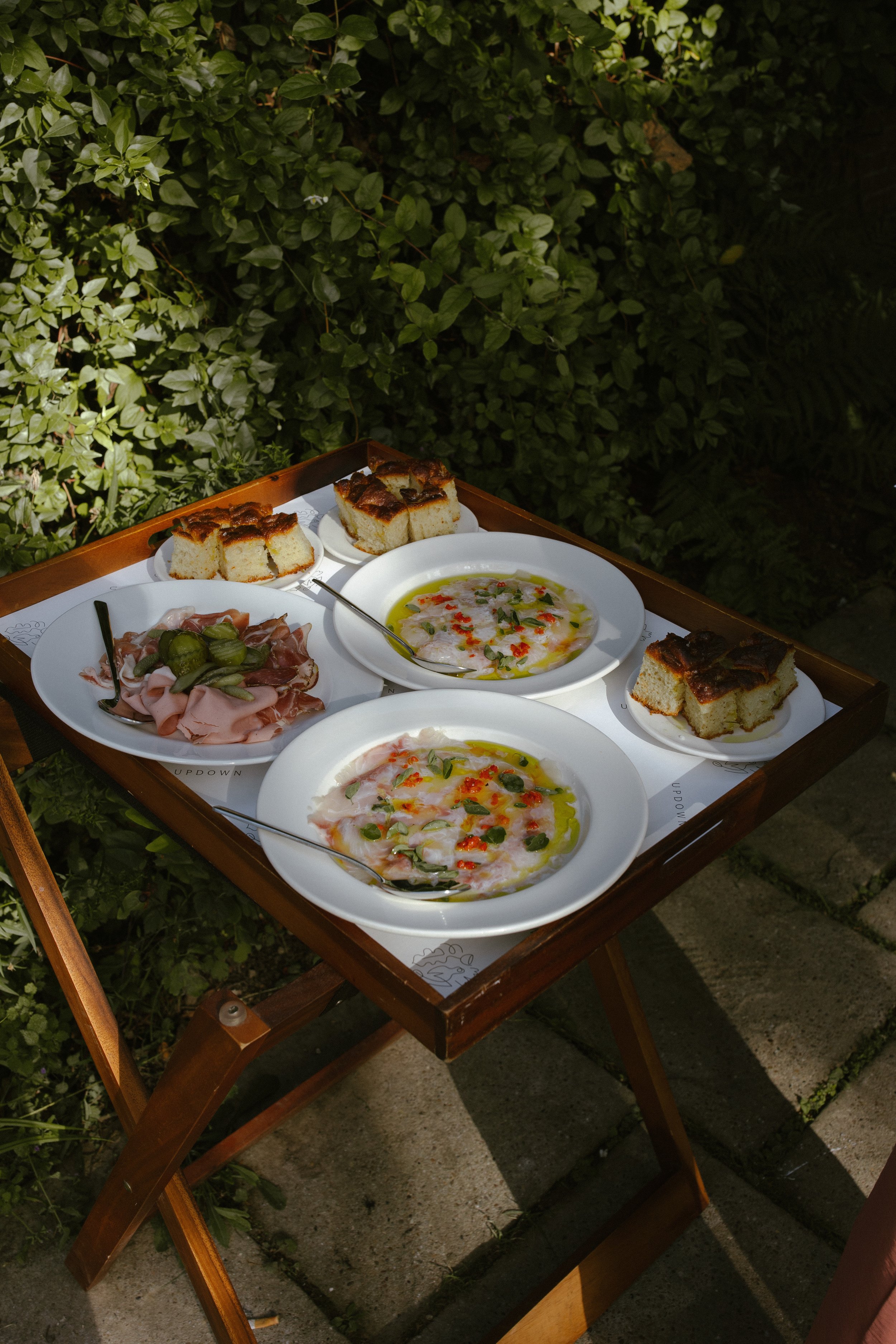 A wooden table with plates of sliced bread, thinly sliced ham with pickles, and two bowls of soup garnished with herbs and chili flakes, set outdoors against green shrubbery.