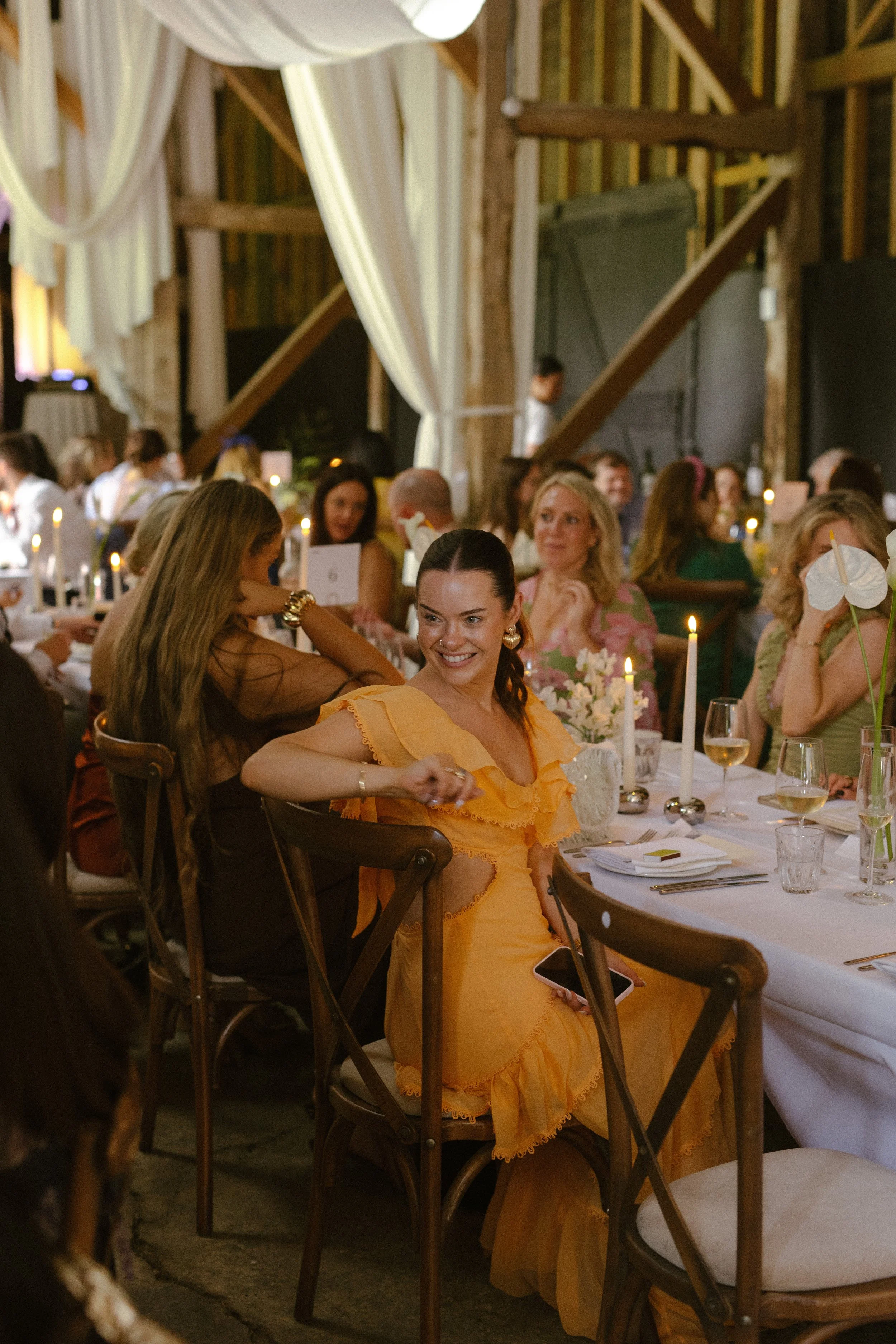 A woman in a yellow dress sitting at a decorated table during a social event, possibly a wedding reception, with other guests around her in a rustic venue with wooden beams and draped curtains.